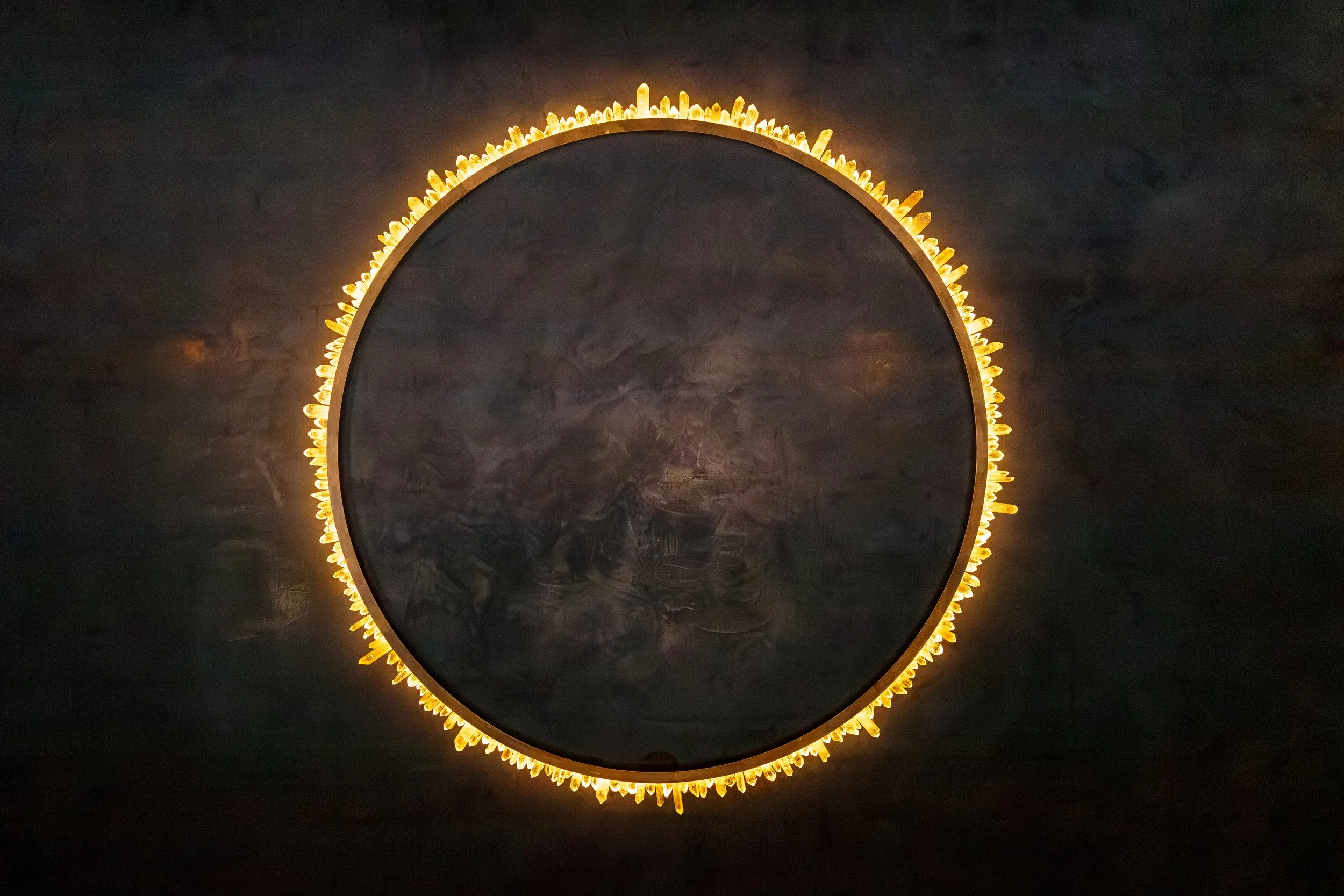 Round mirror with a border of small yellowish crystal-like decorations, mounted on a dark wall.