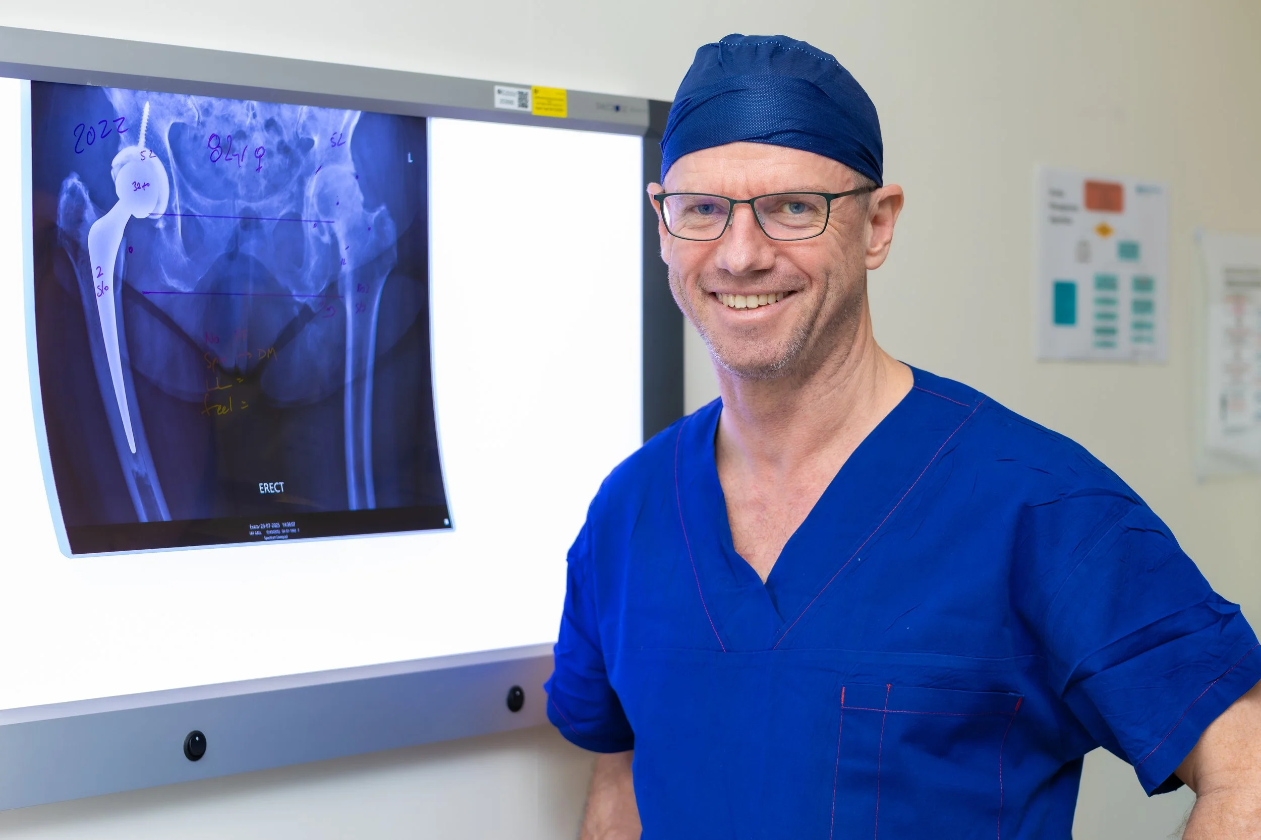 A smiling male medical professional, wearing glasses, blue scrubs, and a blue surgical cap, stands in front of an X-ray display showing a hip joint.