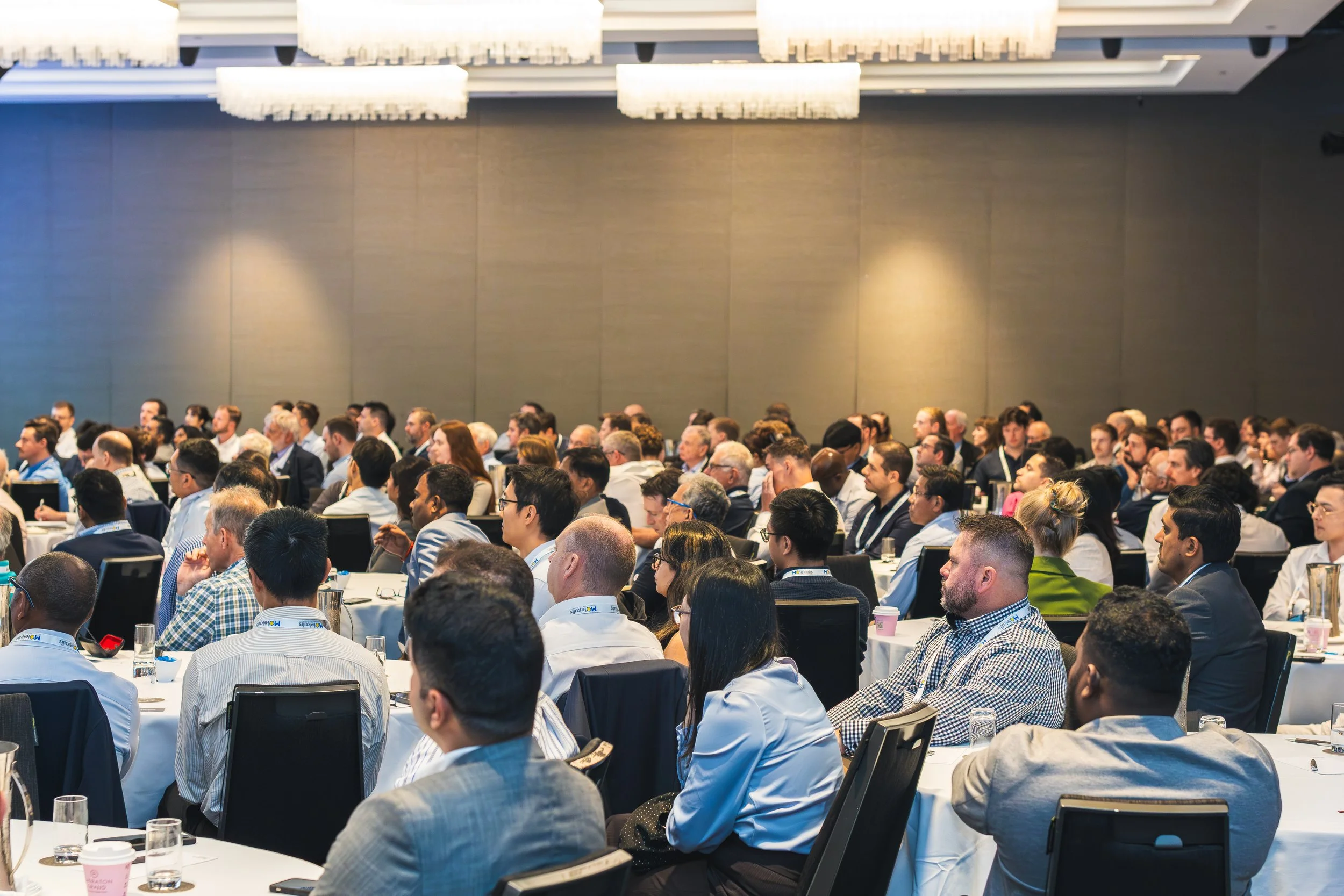 A large conference room filled with diverse attendees seated at round tables, listening to a speaker or presentation at a professional event.
