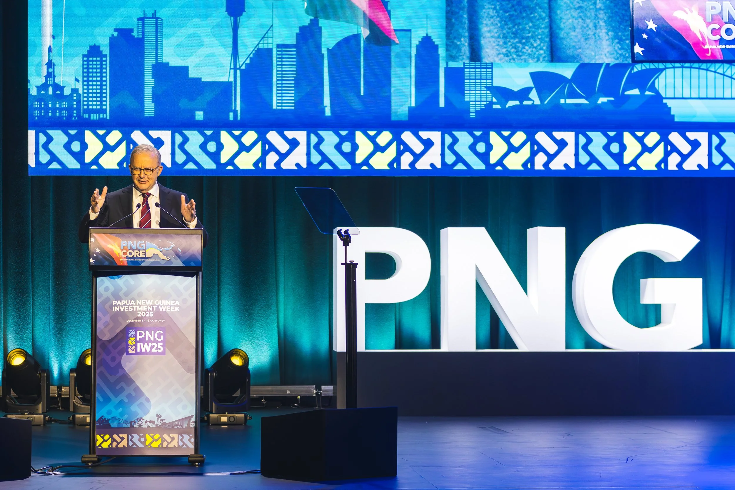 A man in a suit and tie is speaking at a podium during a conference. Behind him is a large screen displaying a blue city skyline with modern skyscrapers, and the event branding for Papua New Guinea Investment Week 2025. The stage is lit with yellow a