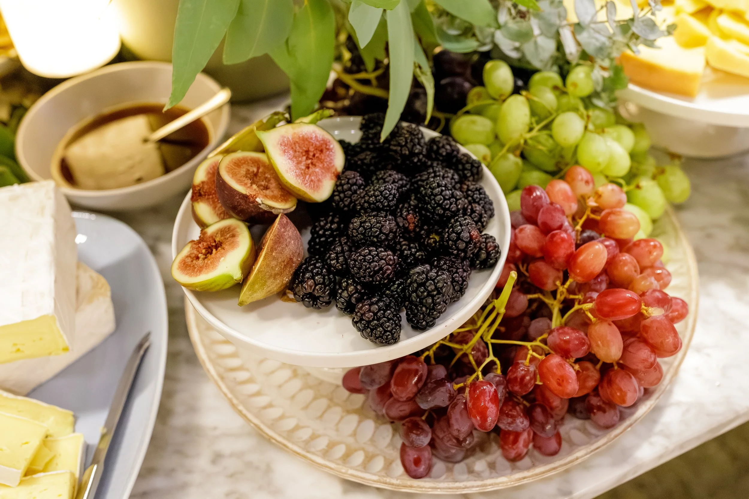 A white bowl of fresh blackberries and sliced figs, surrounded by bunches of green and red grapes, with a plate of cheese and a bowl of caramel sauce in the background.