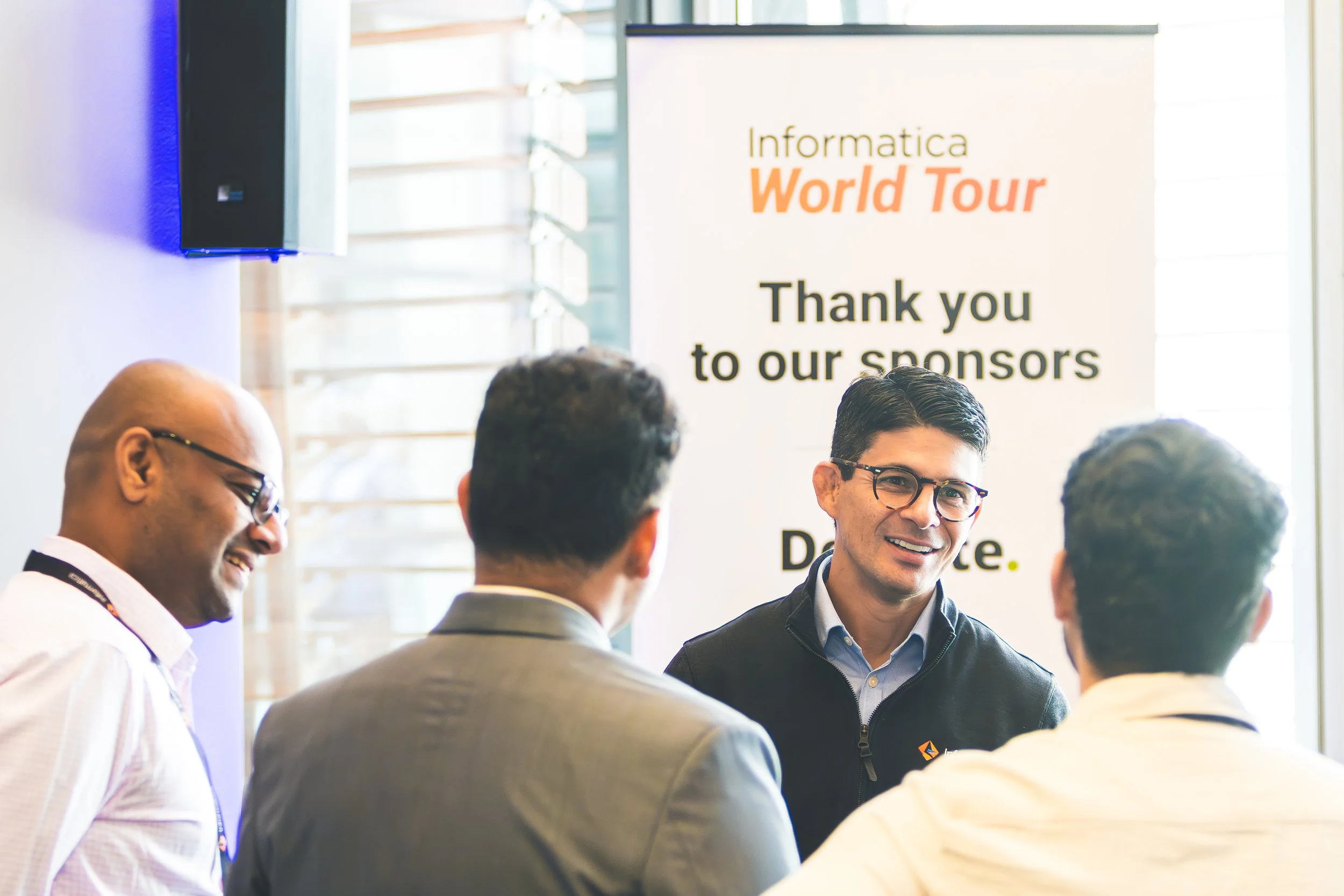 Group of people engaged in conversation at a networking event or conference, with a banner in the background that reads 'Informatica World Tour' and 'Thank you to our sponsors'.