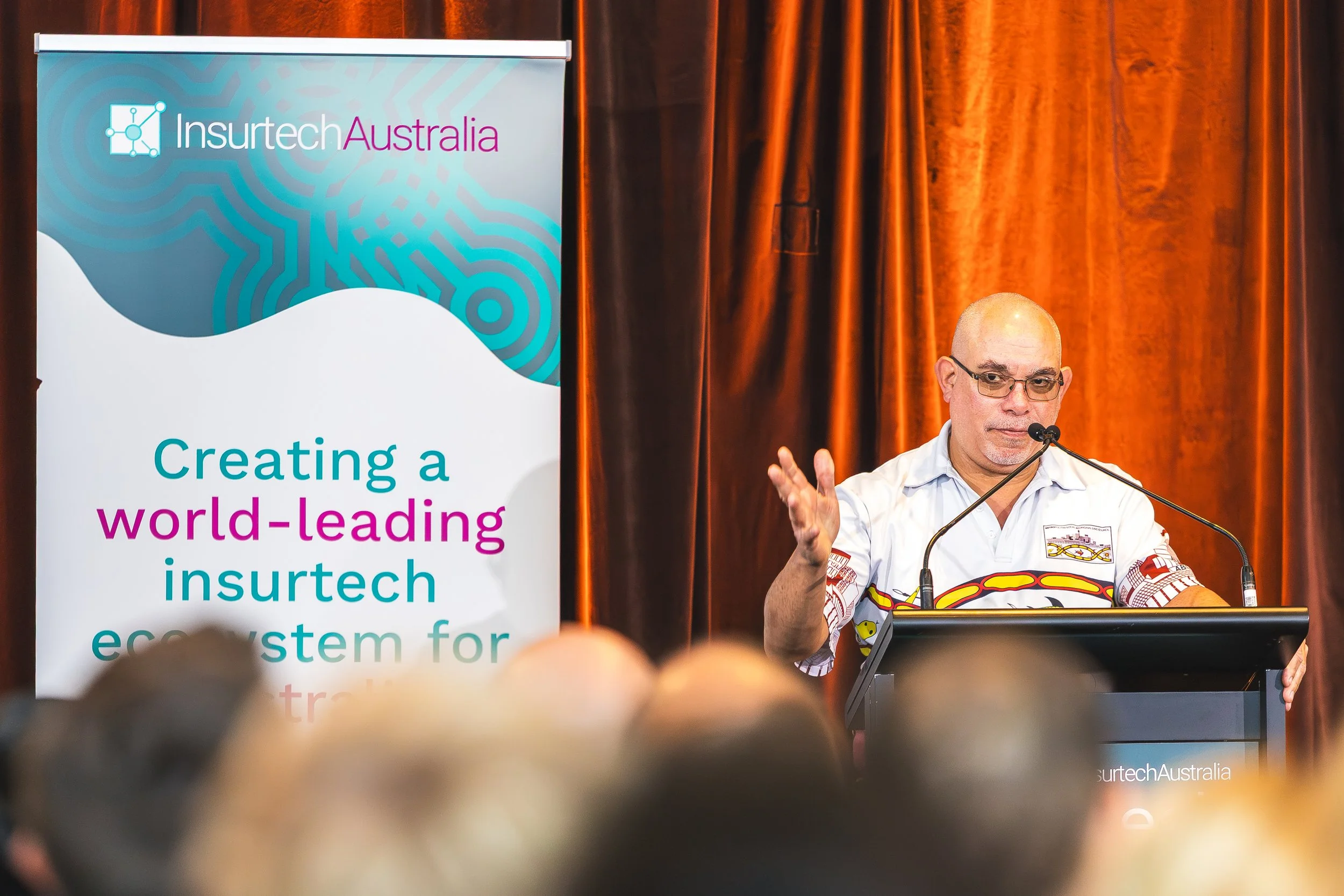 A man wearing glasses and a white shirt with red and yellow patterns, speaking at a podium with a microphone, in front of a large banner for Insurtech Australia. The banner features the text 'Creating a world-leading insurtech ecosystem for...' and h