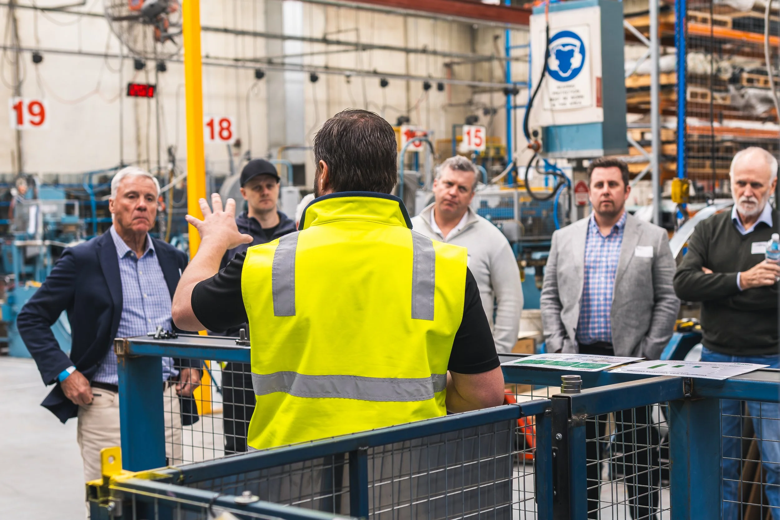 A man with dark hair wearing a yellow safety vest is giving a presentation or tour to a group of five people in an industrial warehouse with machinery and numbered signs in the background.