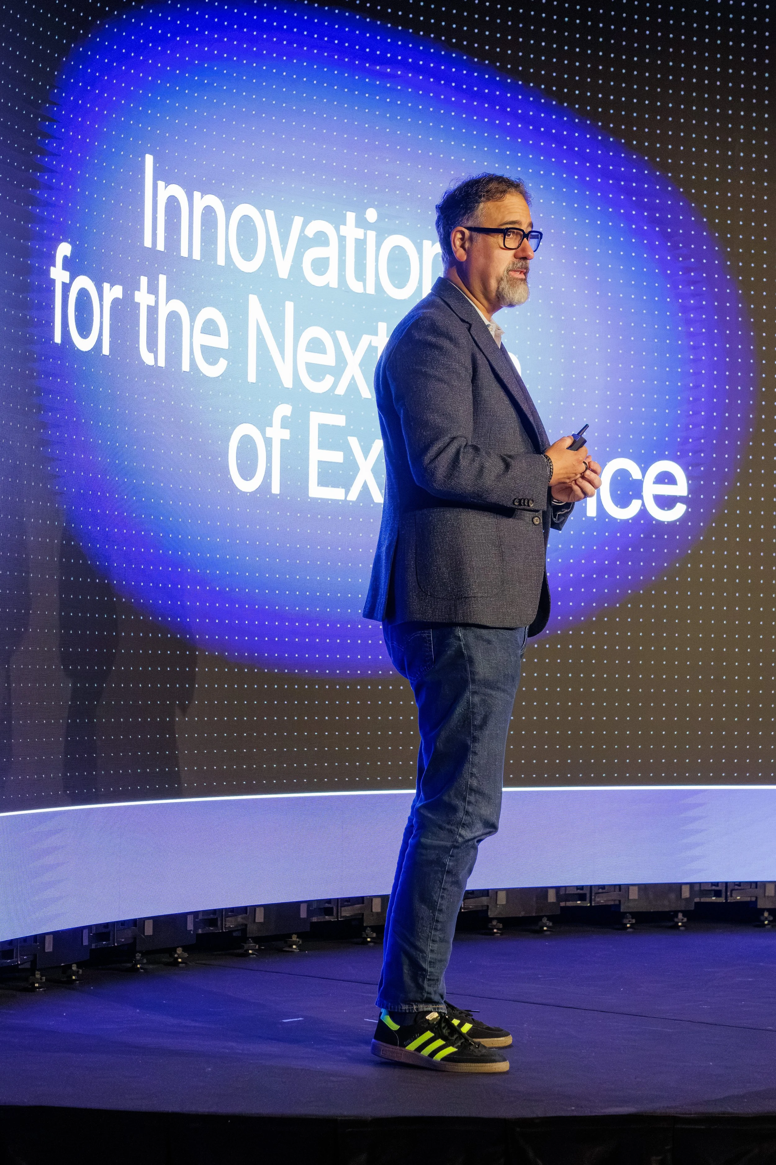 A man in glasses, a gray blazer, jeans, and neon sneakers stands on a stage with a large screen behind him that displays the words 'Innovation for the Next of Experience' in white text on a blue-dotted background.