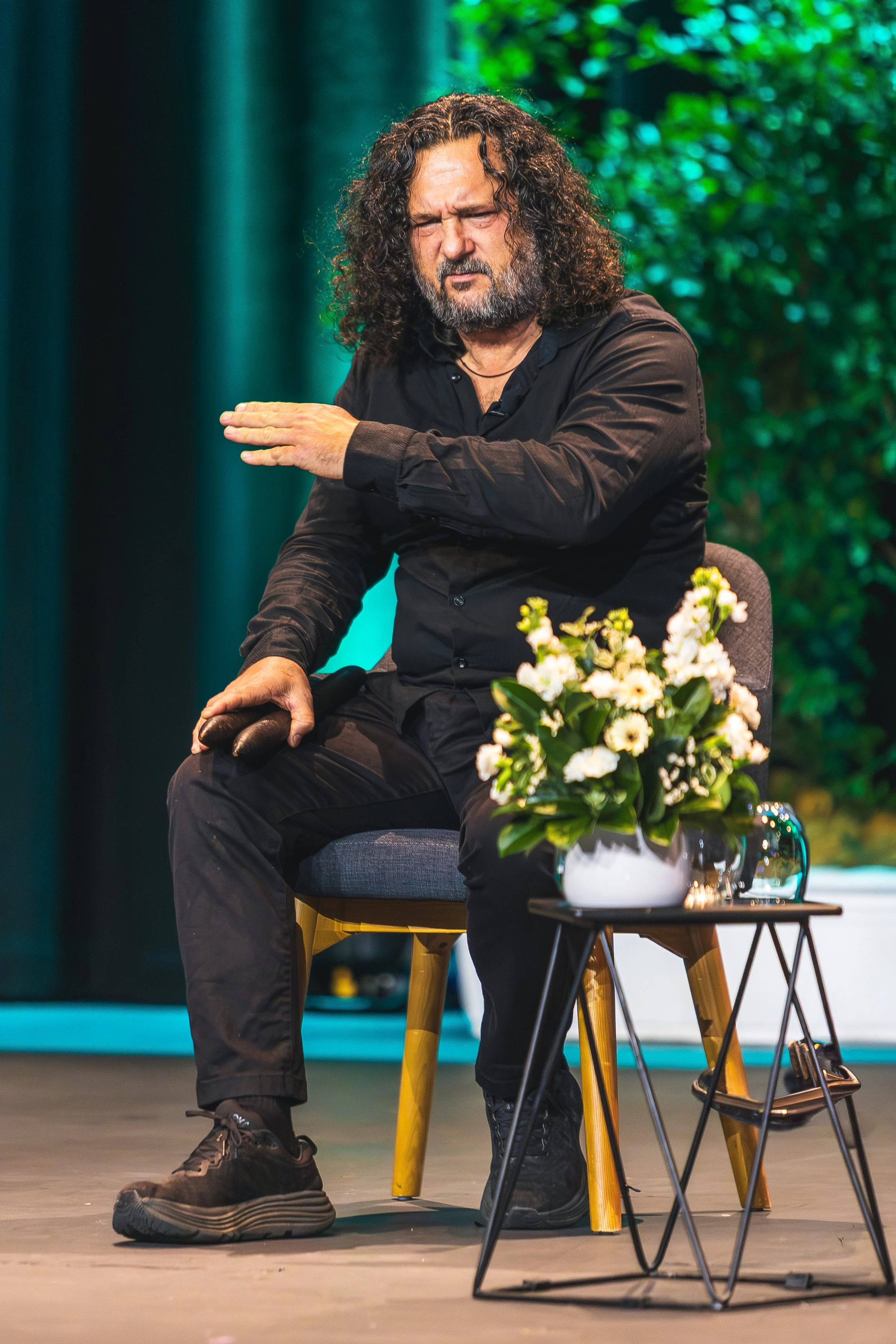 A man with long curly hair, a beard, and dressed in black performs on a stage. He is sitting on a chair with a small table beside him that holds a white vase with flowers. The stage has a green foliage background.