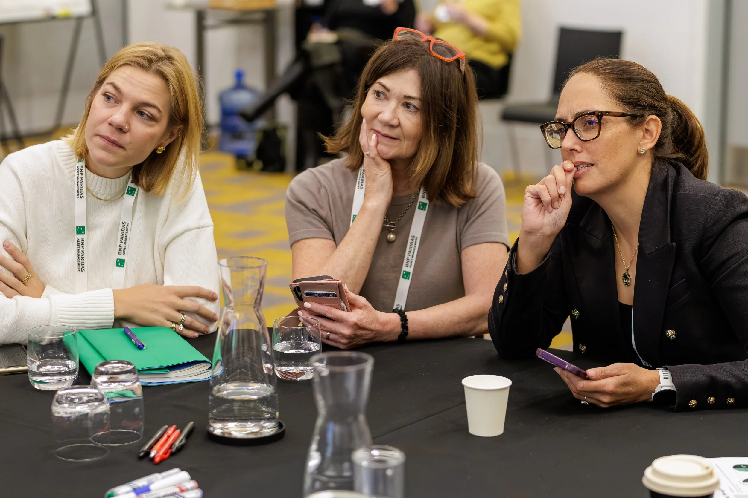 Three women sitting at a black table, engaging in conversation. The woman on the left has blonde hair, wearing a white sweater and earrings. The woman in the middle has brown hair, orange sunglasses on her head, and is holding a phone. The woman on t