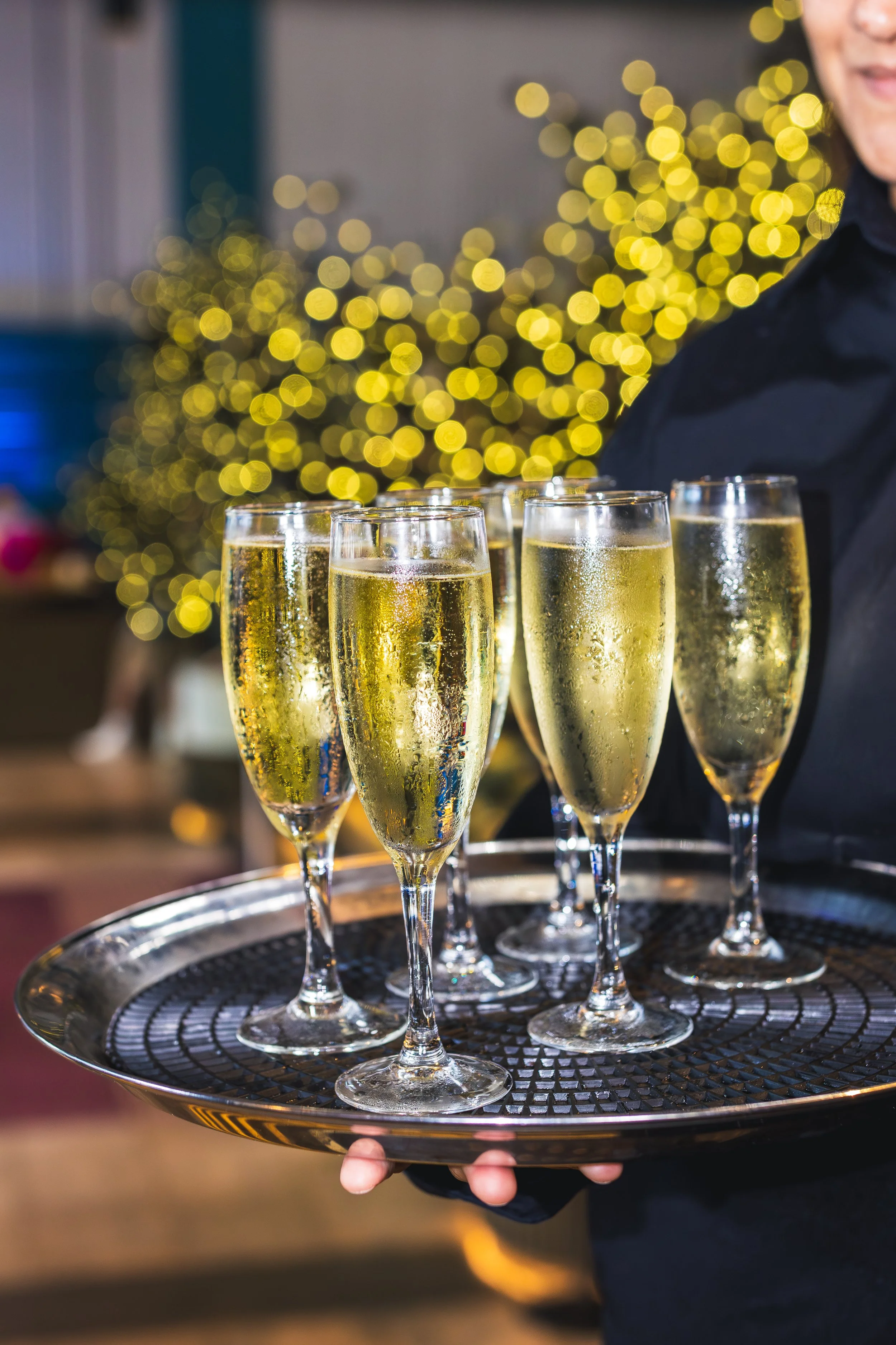 Tray of six glasses of champagne with bubbles, held by a person in black attire, against a blurred background of golden lights and a person's smile.