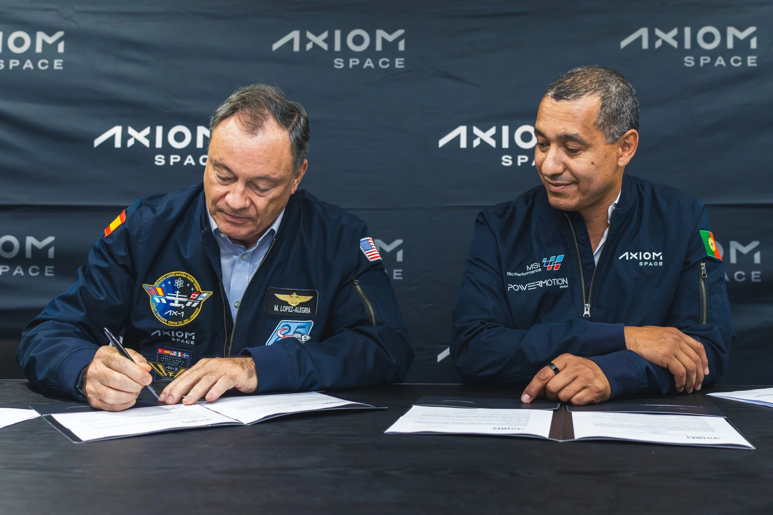 Two men in space agency jackets signing documents at a table, with a backdrop displaying the 'Axiom Space' logo.