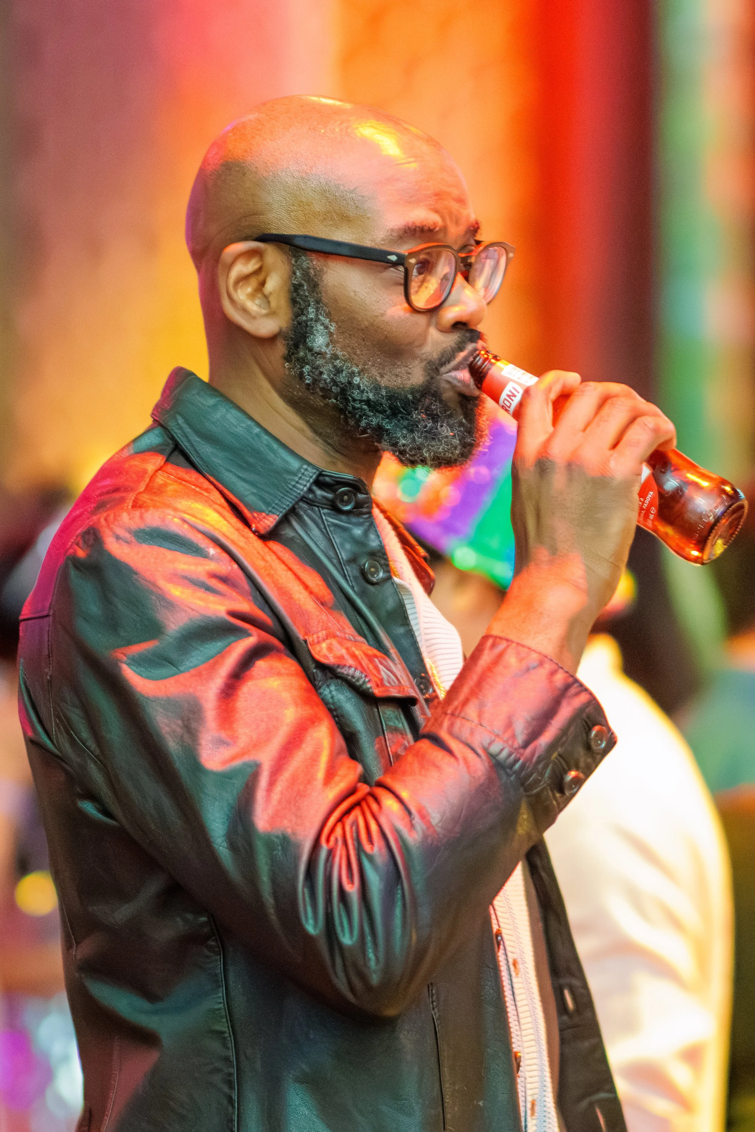 A man with a bald head, glasses, and a gray beard drinking from a brown bottle, wearing a black and red jacket with a striped shirt underneath.