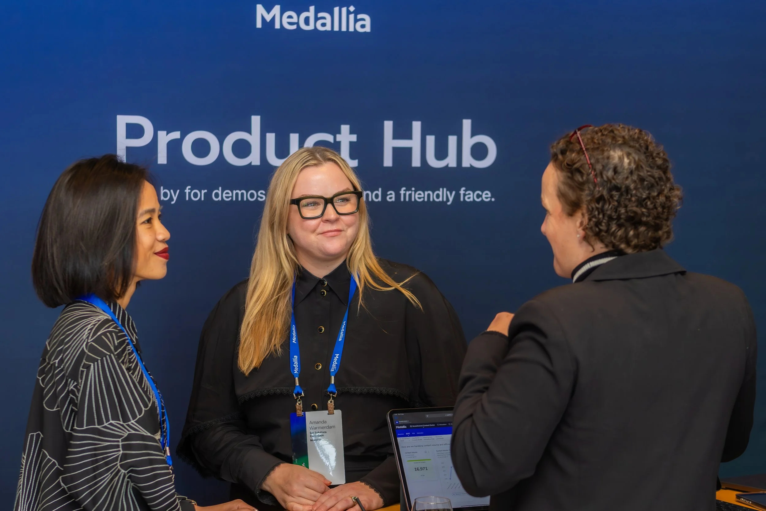 Three women engaged in a conversation at a Medallia event, with a large blue Medallia Product Hub sign in the background.