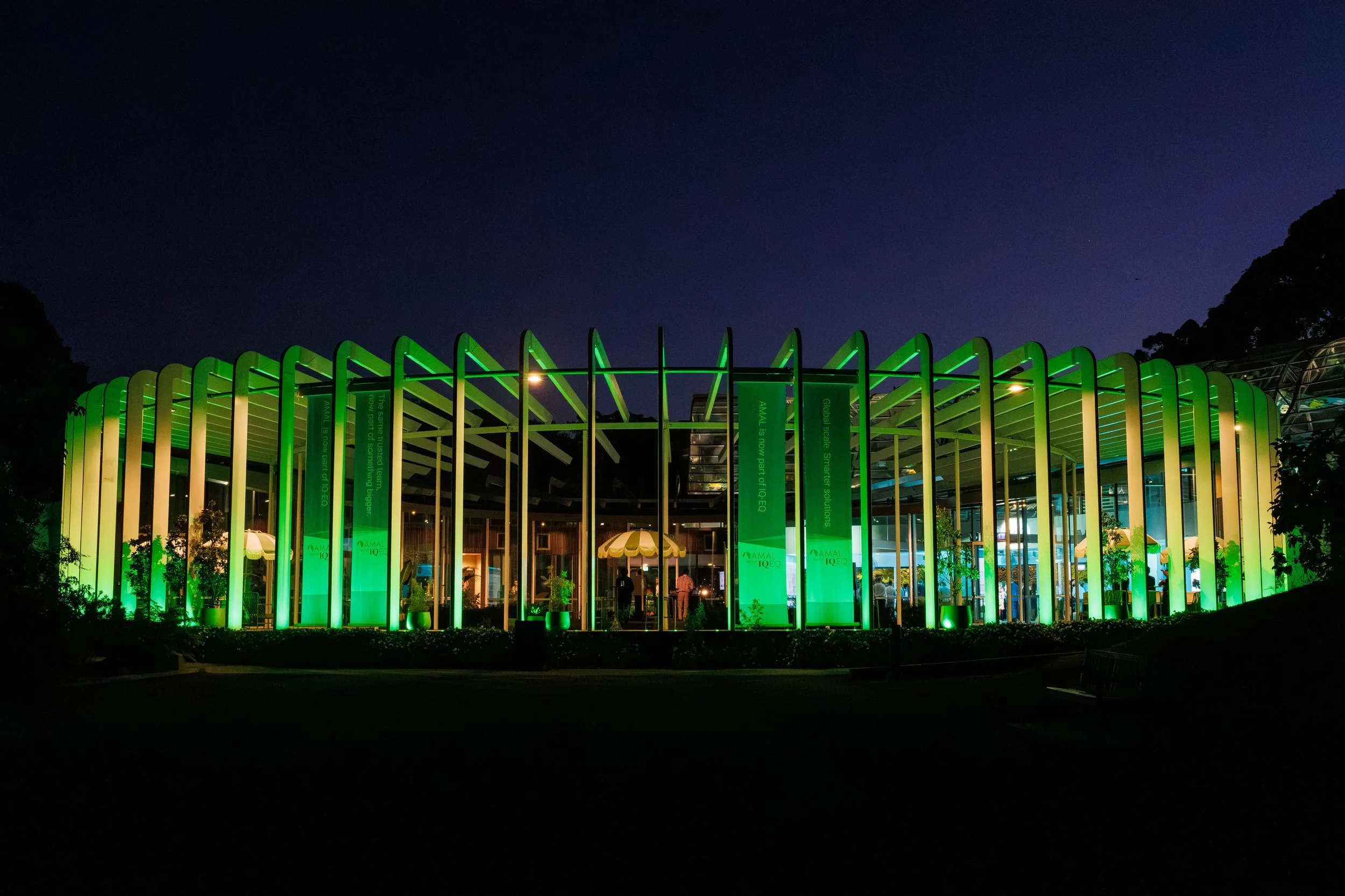 Night view of a modern building with green illuminated vertical pillars and a dark blue sky.