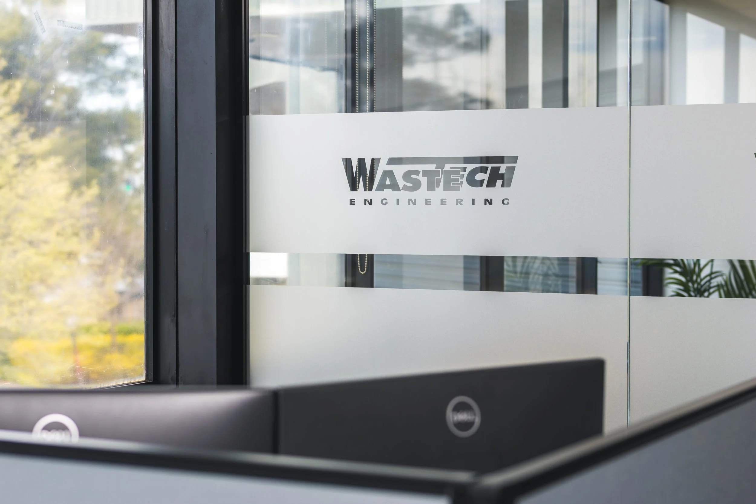 Close-up of a glass door with the logo 'WASTECH ENGINEERING' on it, with reflections of trees and buildings outside.