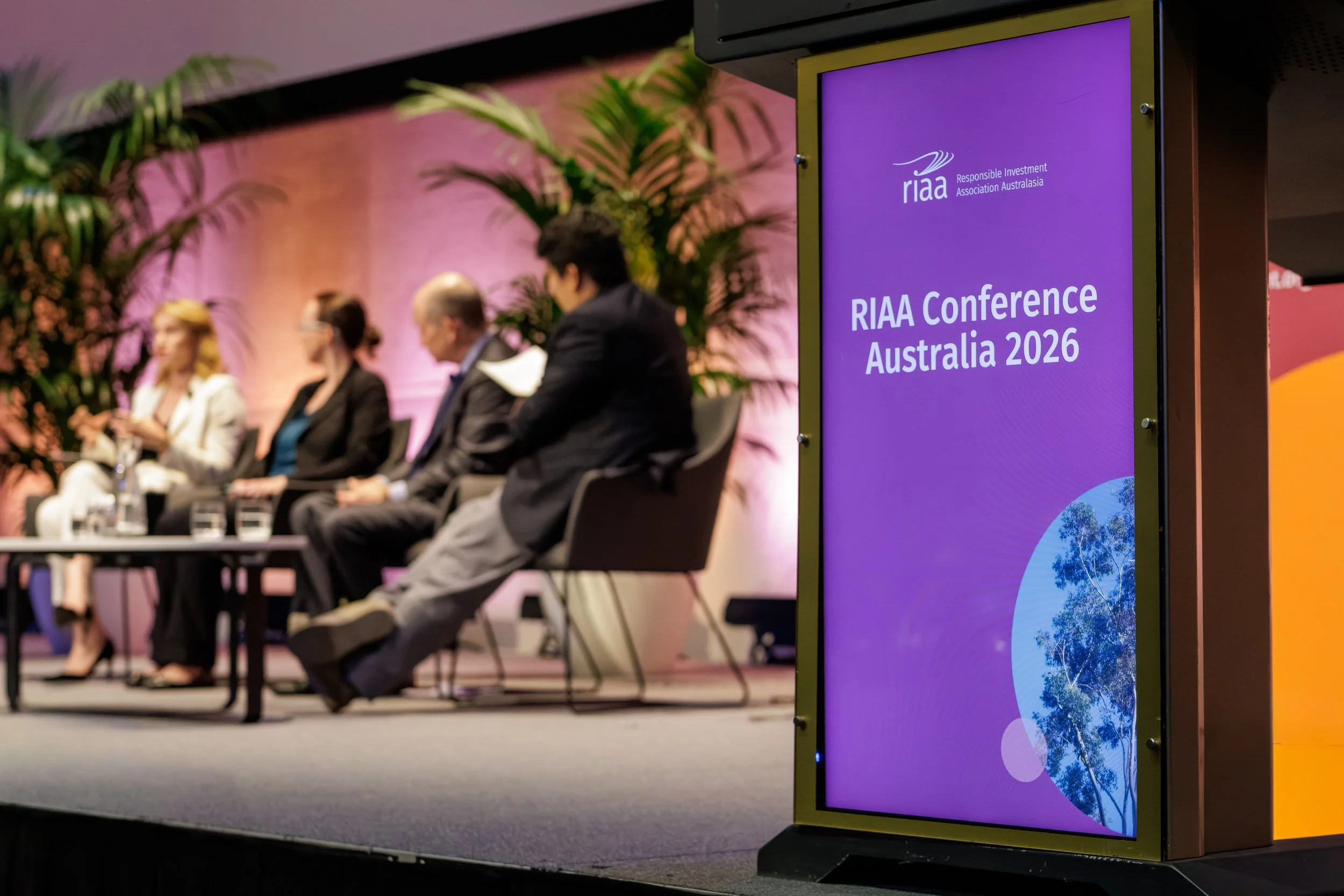 Panel display at the RIAA Conference Australia 2026, showing a purple background with the conference name and logo, with blurred people sitting on stage in the background.