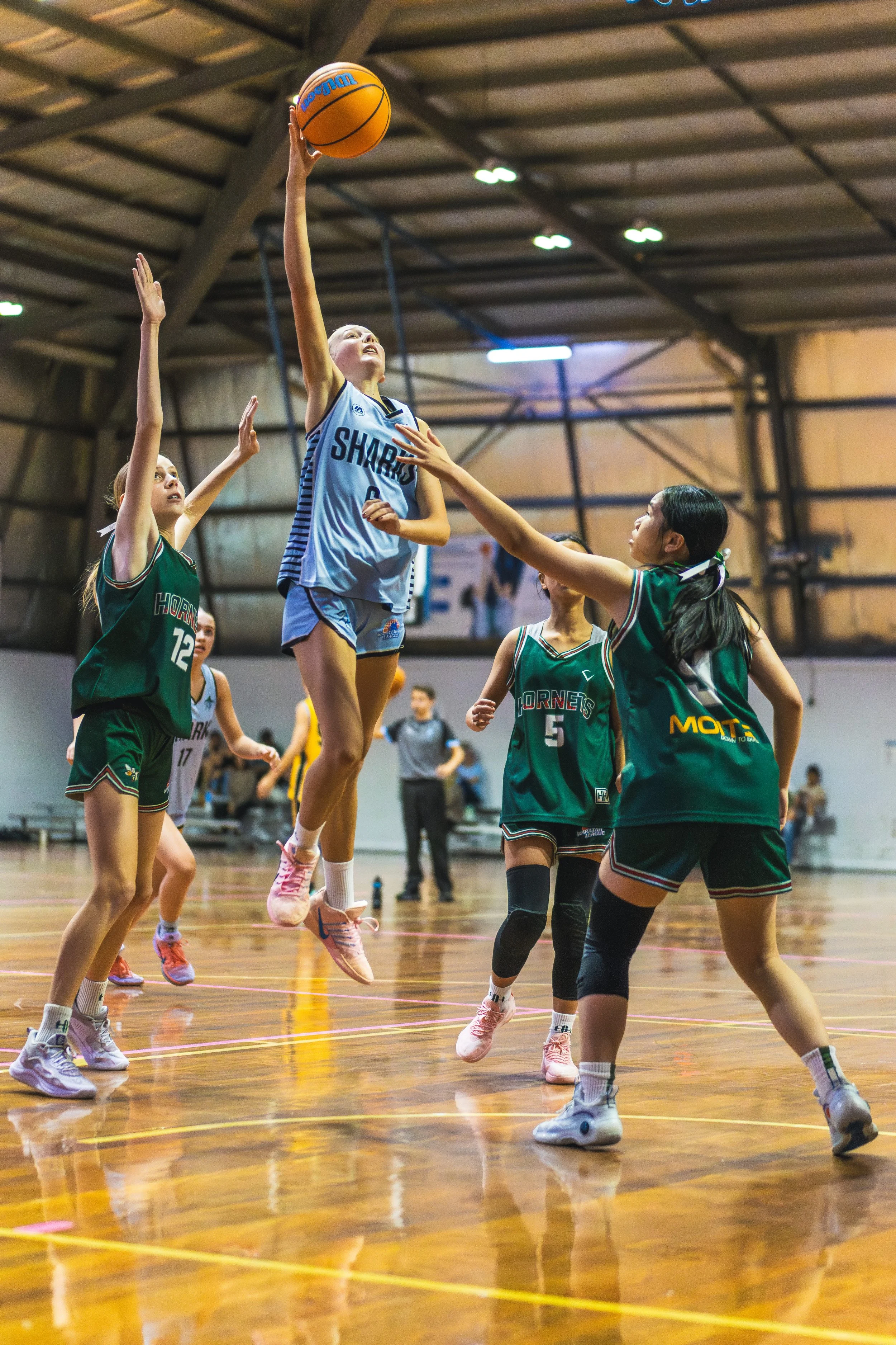 Young female basketball player in white uniform jumping to make a shot during a game, with opposing players in green defending.