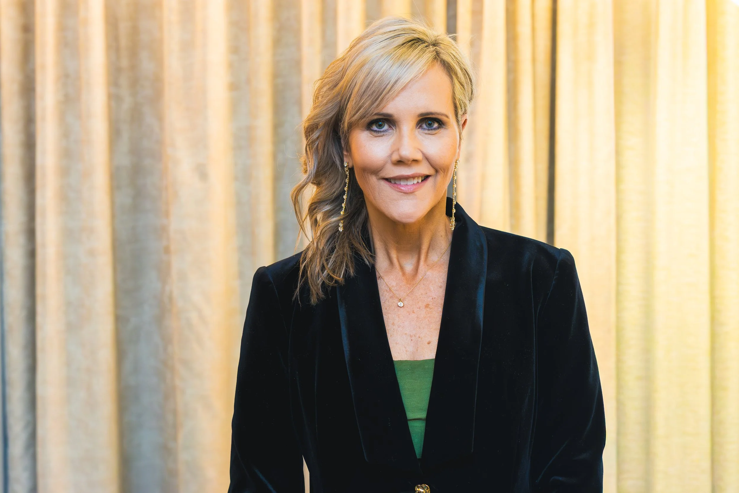 A woman with blonde hair and blue eyes smiling, wearing earrings, a black blazer, and a green top, standing in front of a beige curtain.