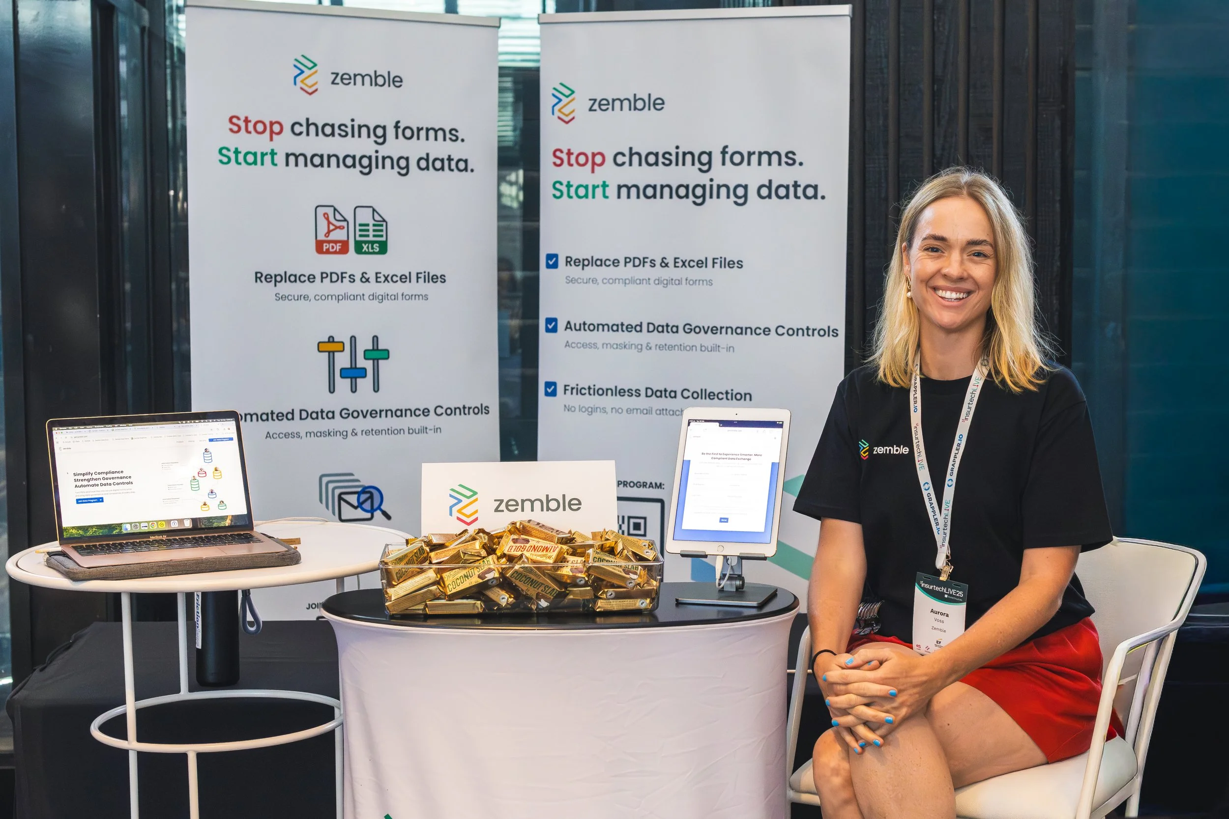 A woman smiling at a booth for Zembla, with posters displaying data management services behind her, a laptop, a tablet, and a bowl of snack bars on the table.