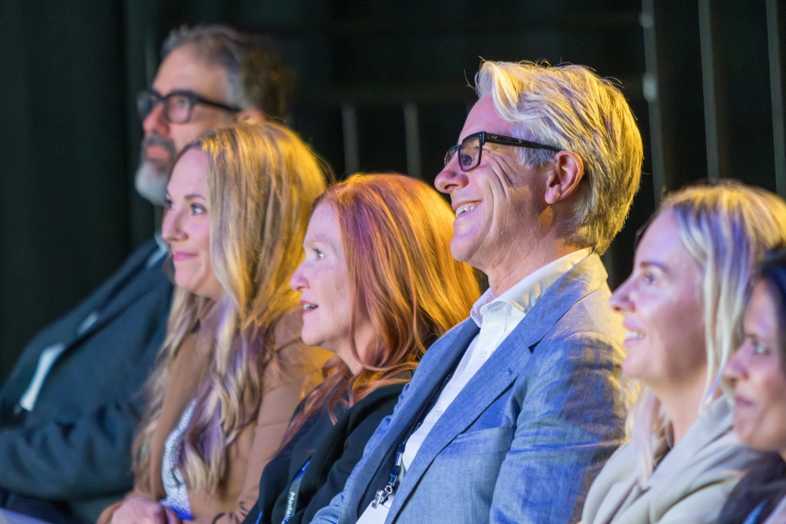 A group of people sitting in a row at a conference, smiling and listening attentively.