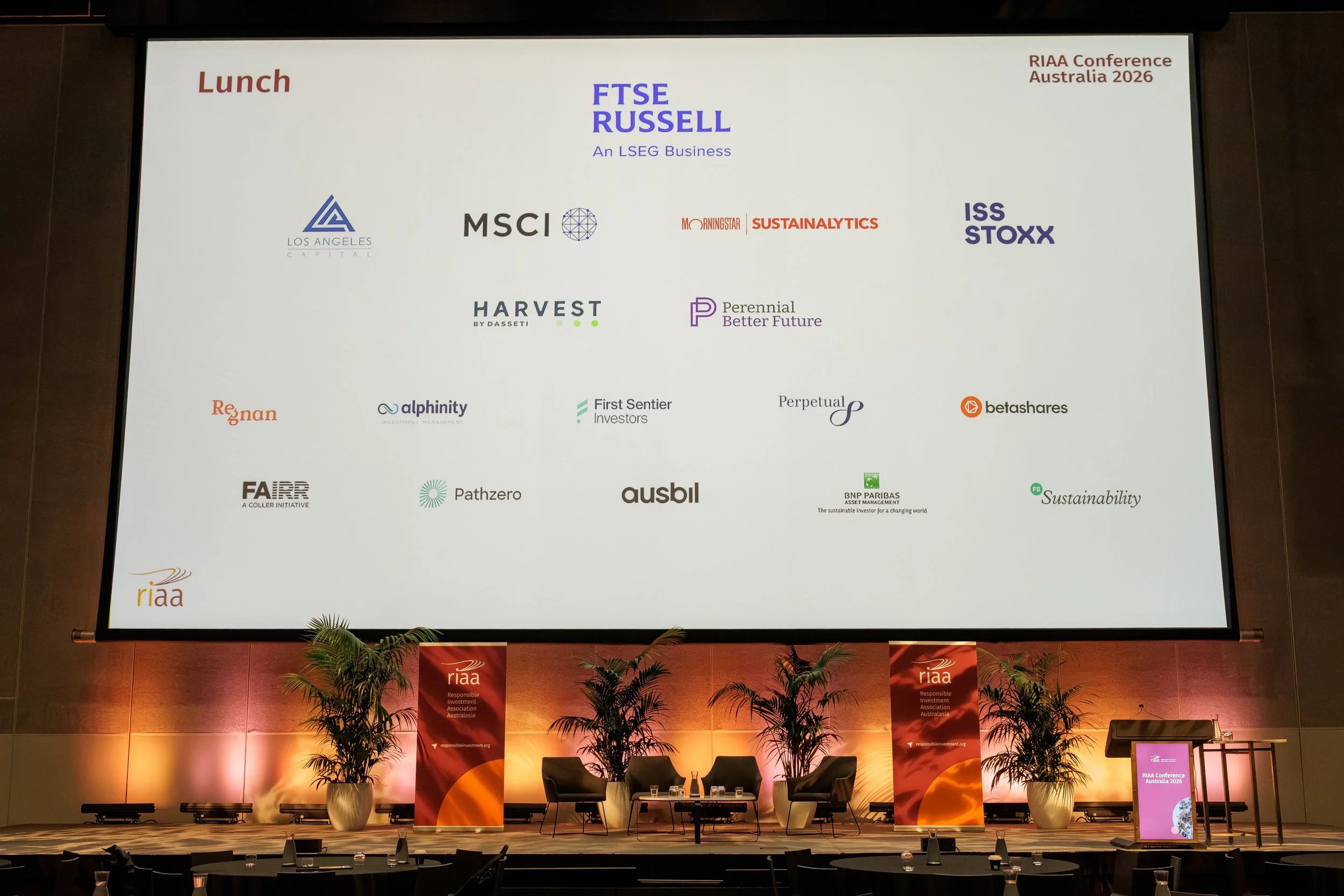Large conference stage with a big screen displaying the logo of RIAA Conference Australia 2026 and the names and logos of various companies and organizations. The stage has chairs, cups, and potted plants, with banners and tables at the front.