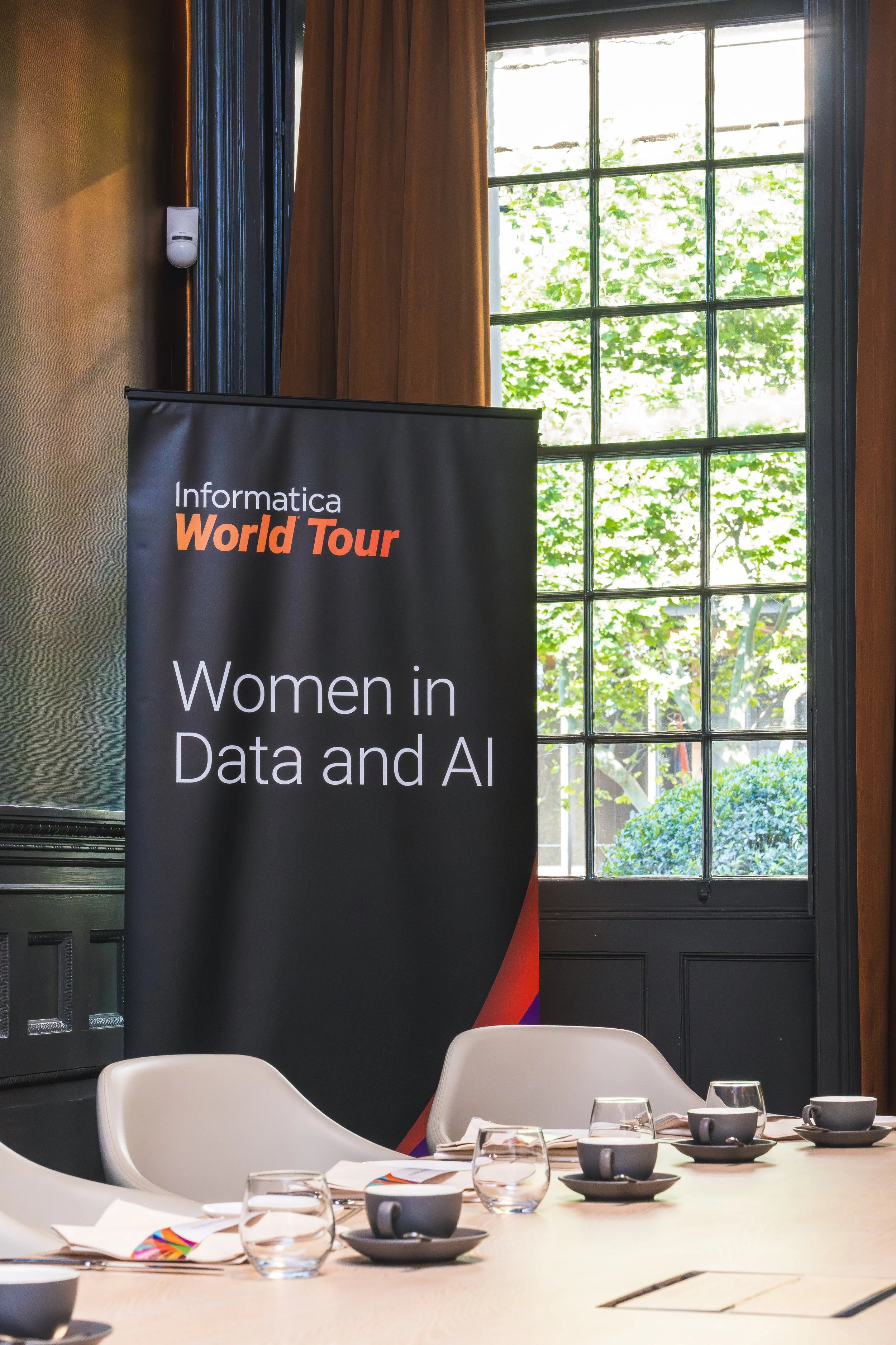 Conference room with a large window and green trees outside. There's a black banner with orange and white text that reads 'Informatica World Tour' and 'Women in Data and AI'. The table has white chairs, glasses, cups, and napkins.