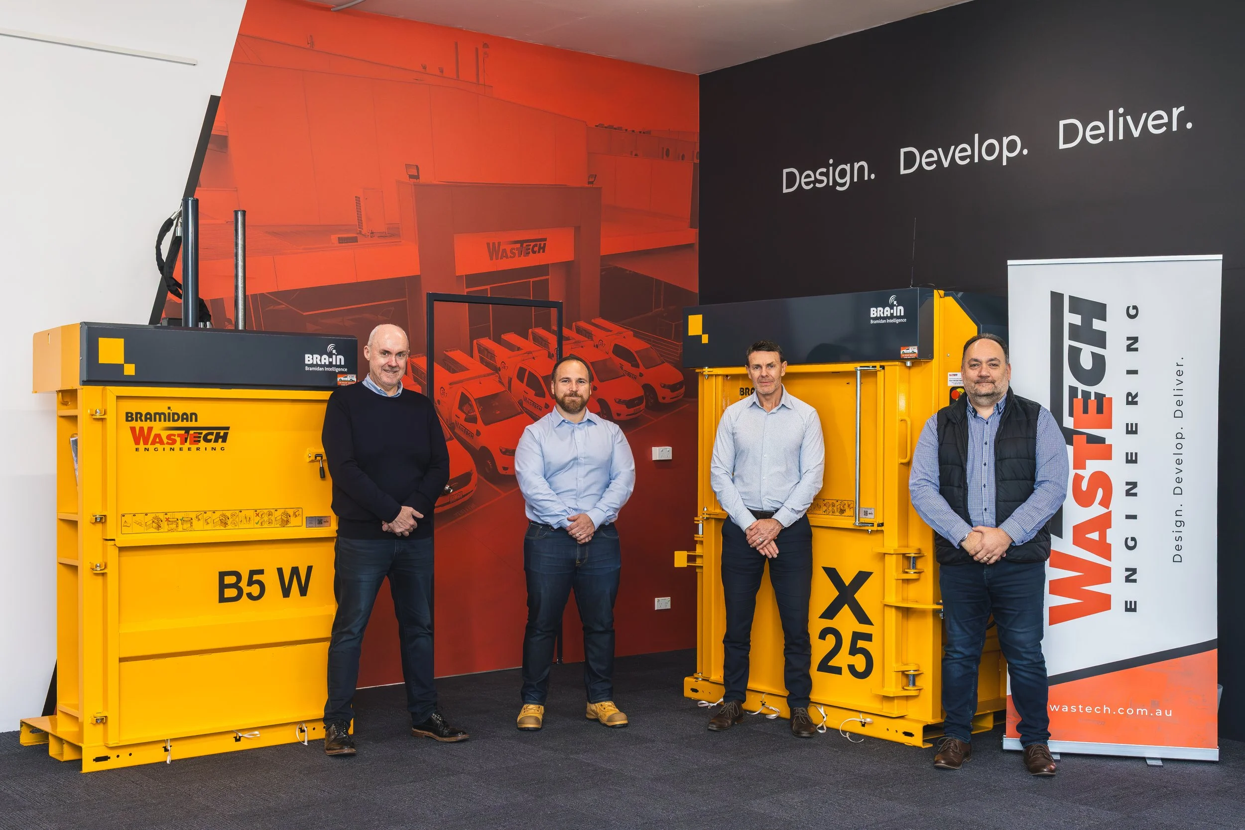 Four men stand in front of large yellow waste management containers labeled B5 W and XG 25, with a red and black Wastech banner to the right. The background features a mural of vehicles and a building with the Wastech logo, and a black wall with the 