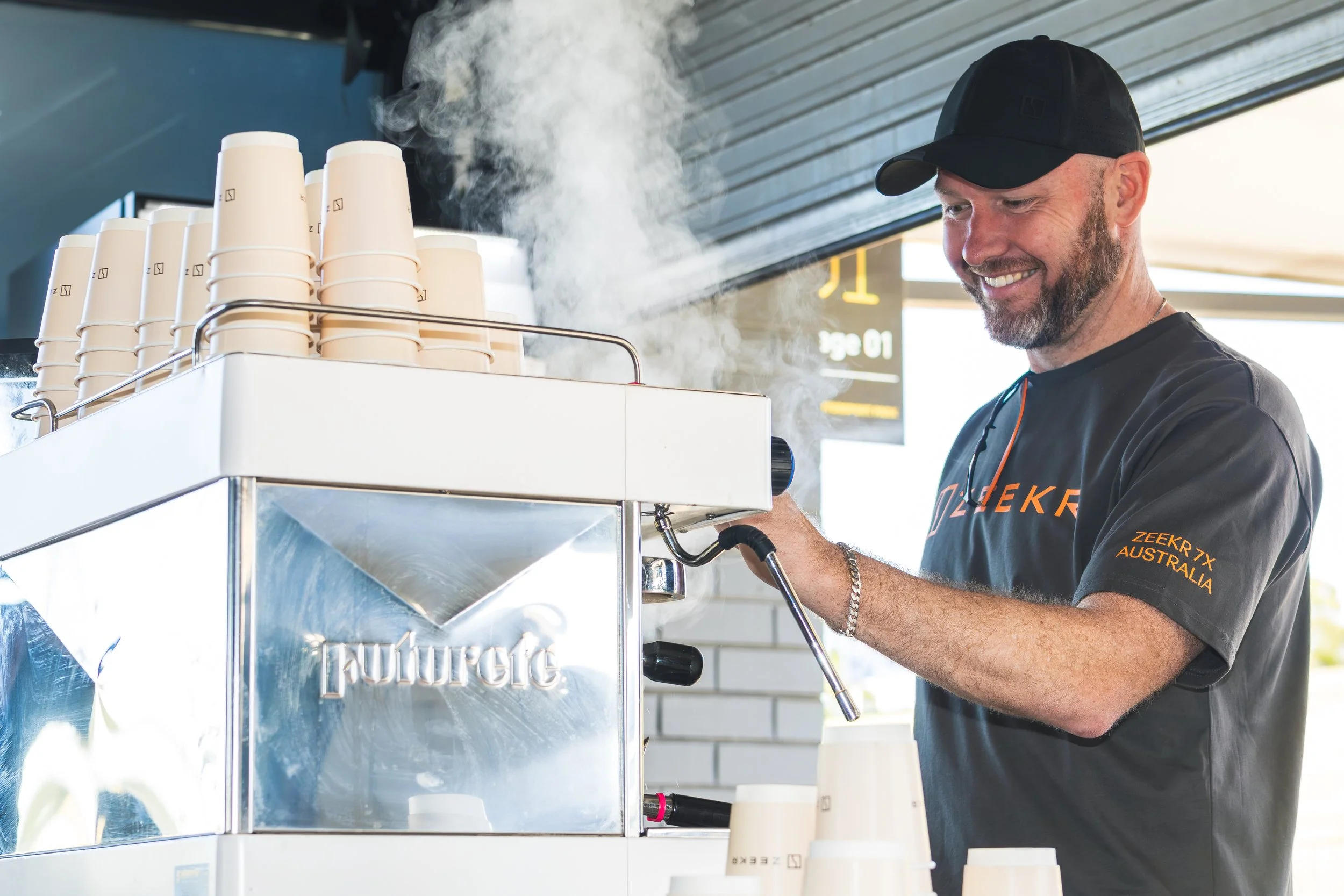 A man wearing a black cap and gray T-shirt smiling while making coffee on a commercial coffee machine inside a cafe or coffee shop.