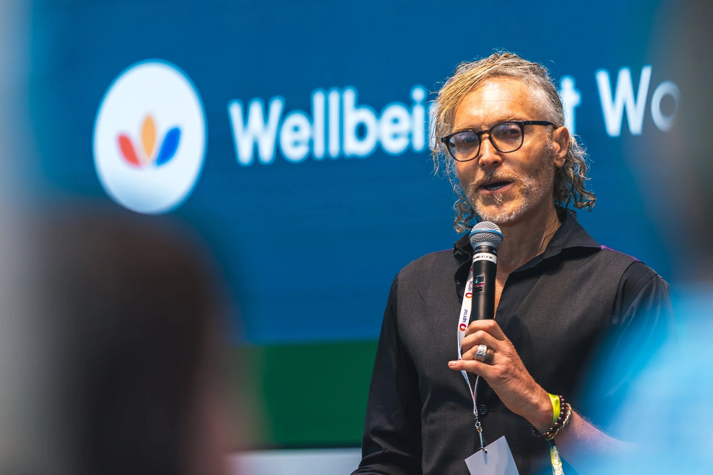 A man with gray hair and glasses speaking into a microphone at an event with a blue backdrop that has the words 'Wellbeing' and a logo.
