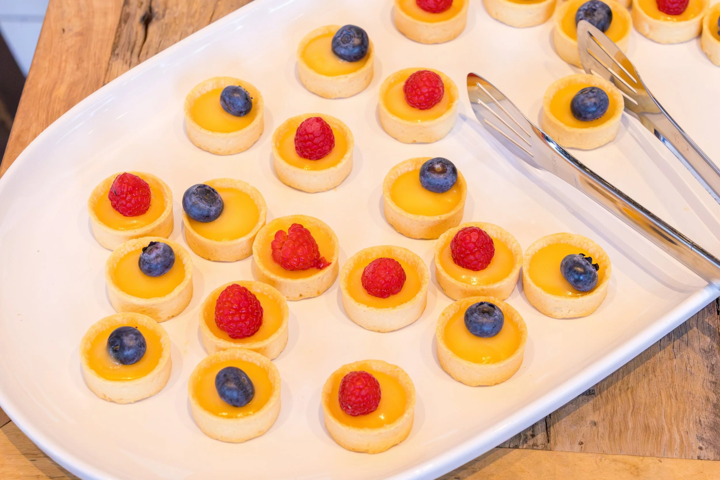 A white rectangular platter with small lemon tartlets topped with blueberries and raspberries, arranged in neat rows on a wooden table.