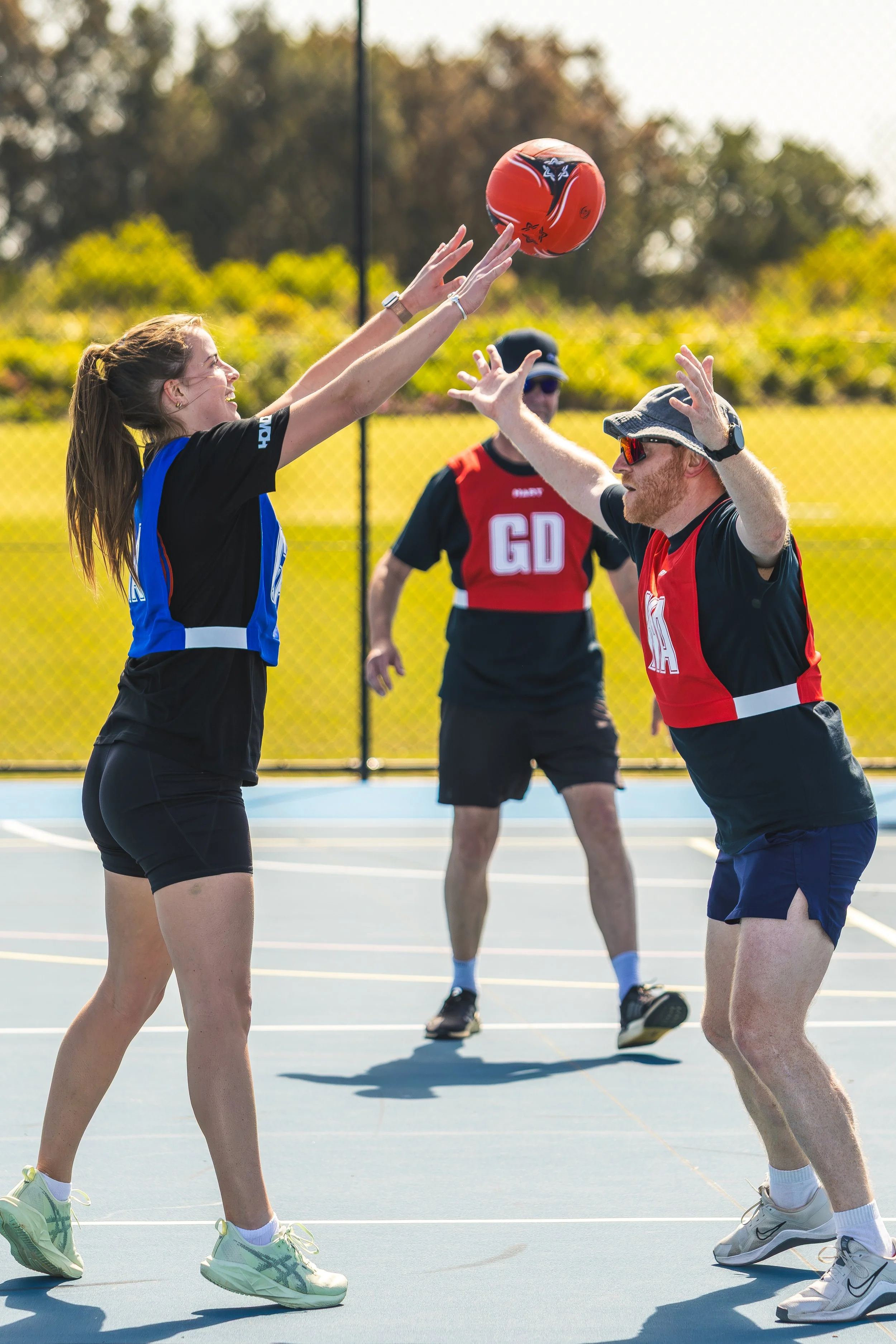 Young woman and man playing basketball on an outdoor court, with a referee or coach watching in the background during bright, sunny weather.