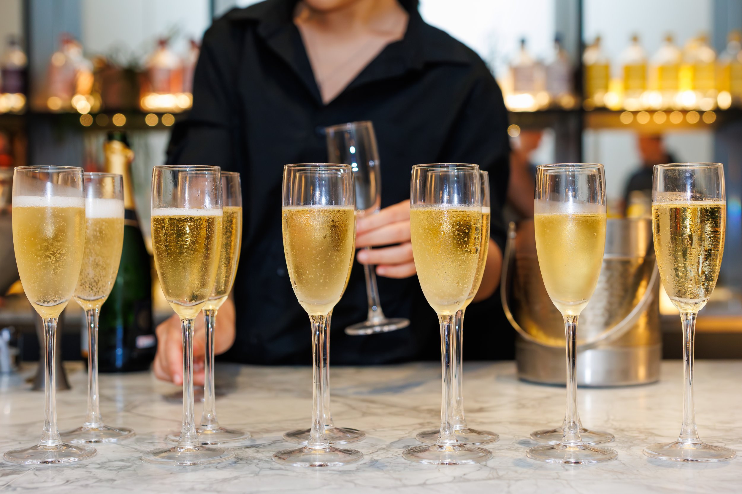 A bartender holding a wine glass of champagne behind a row of filled champagne flutes on a marble countertop.
