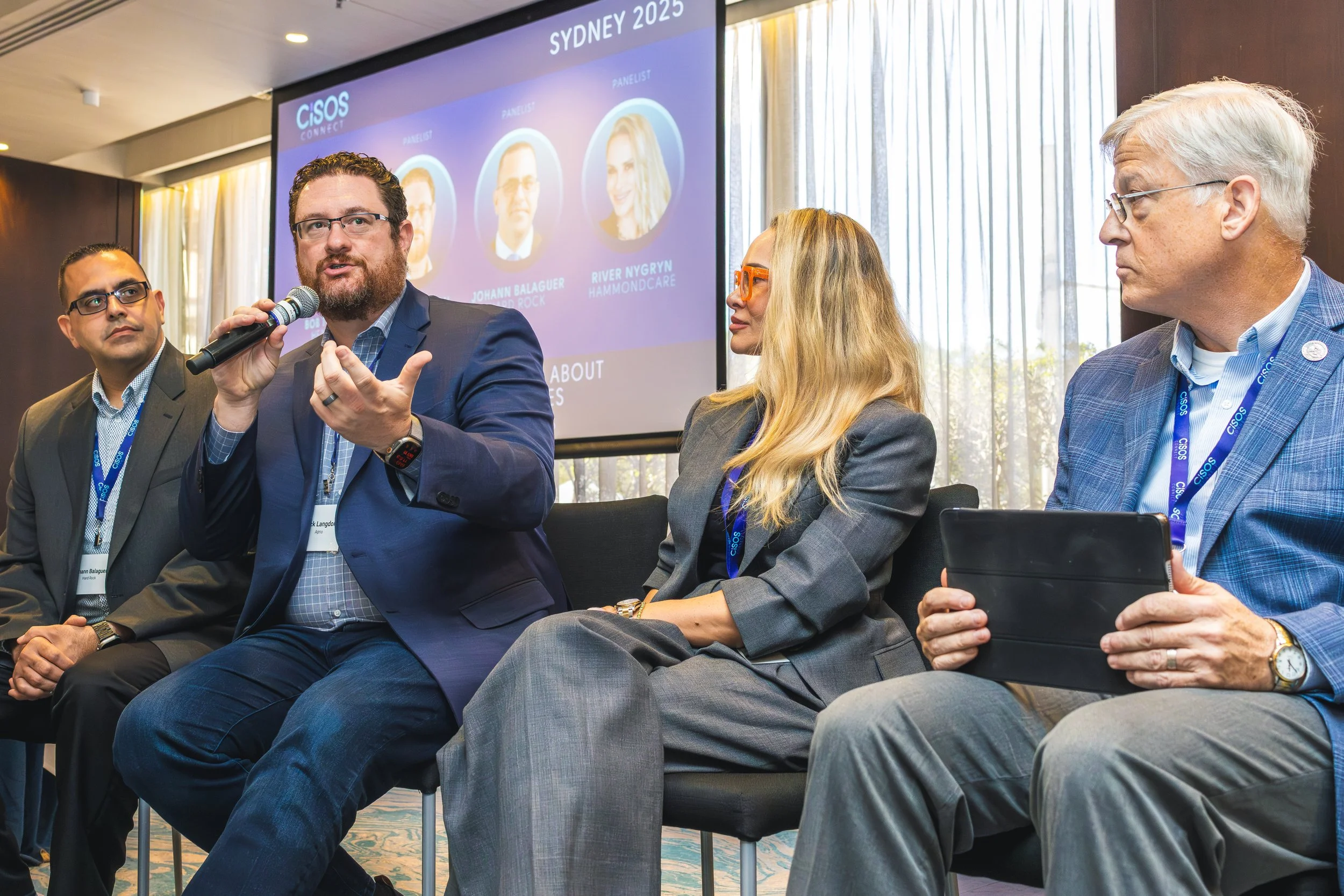 Four people sitting at a conference panel in front of a large screen, with the man in the center speaking into a microphone. The screen displays the event title 'SYDNEY 2025' and images of panelists. The scene is indoors with natural light coming thr