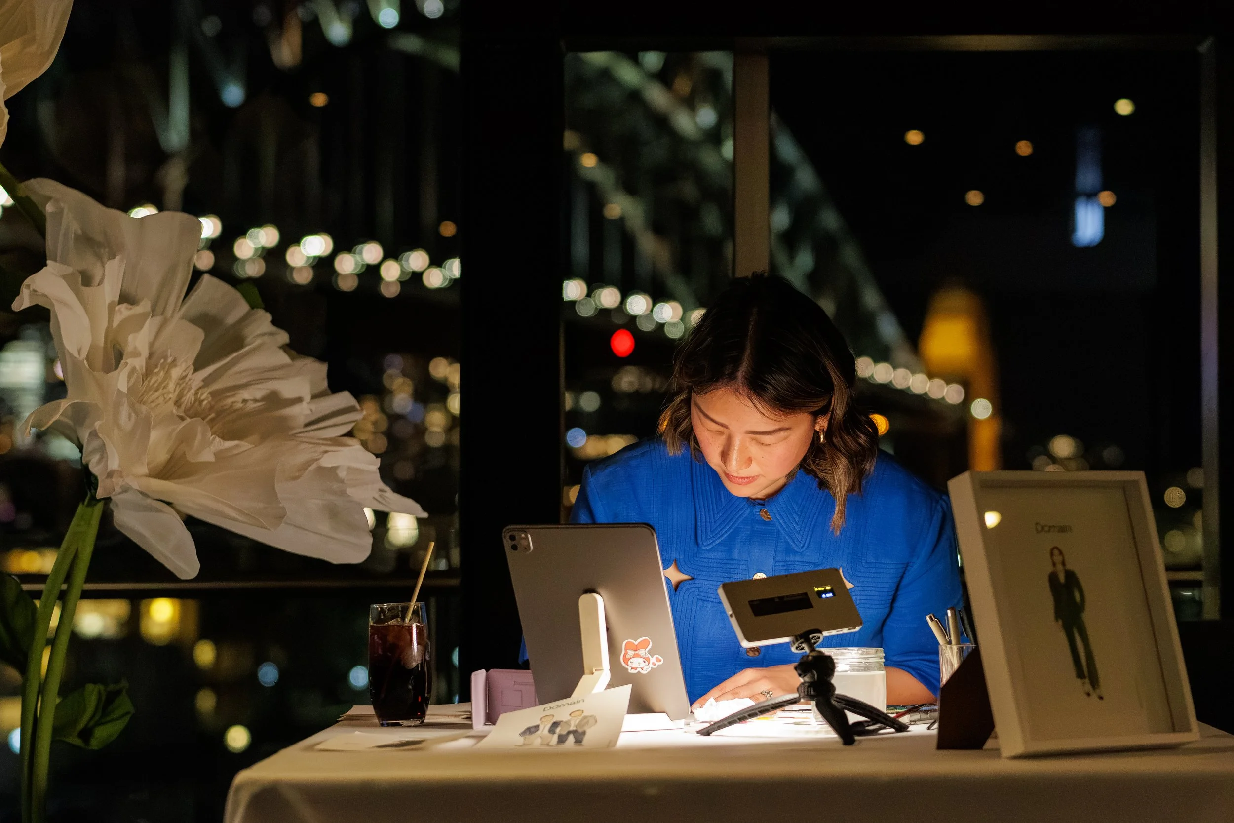 A woman wearing a blue shirt working at a restaurant counter with electronic devices, flowers, and a drink in front of her at night, with blurred city lights in the background.