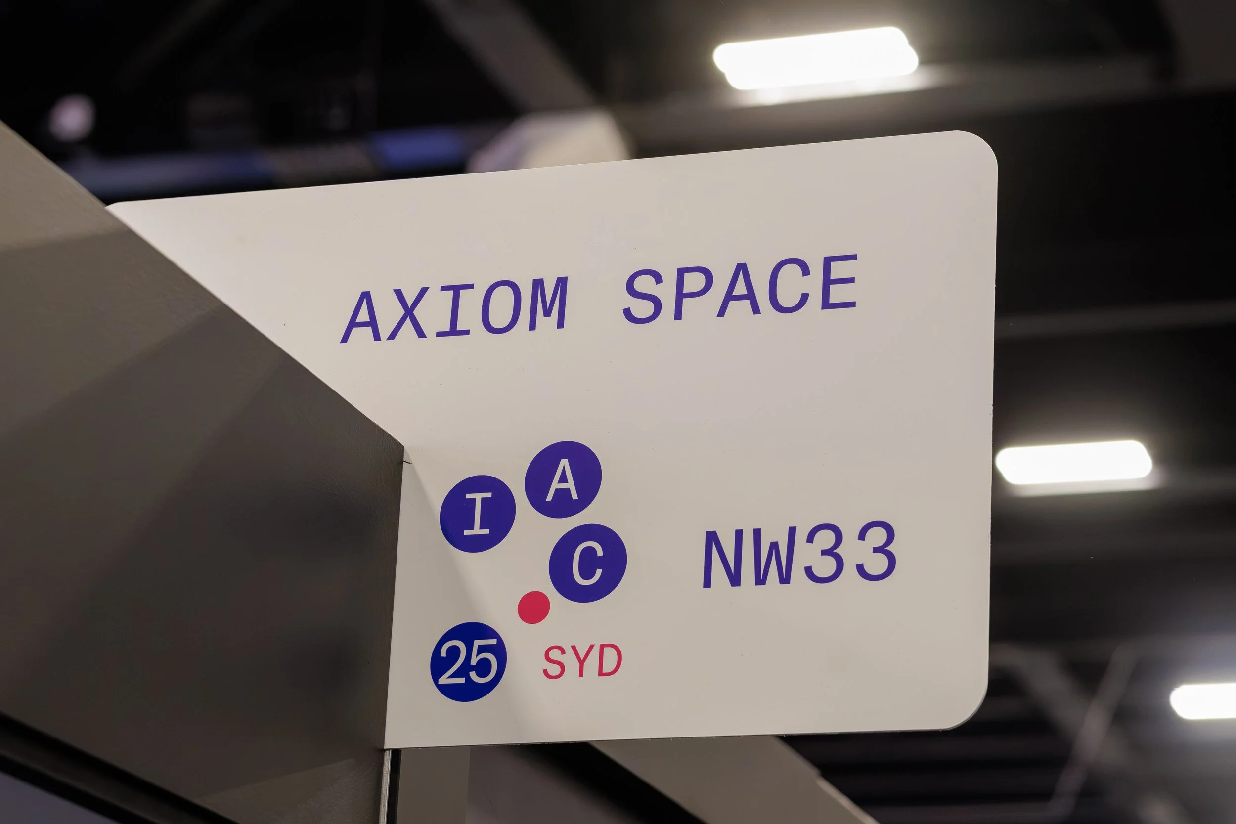 A sign with the text 'AXIOM SPACE' and circle icons with letters and numbers, displayed indoors under ceiling lights.