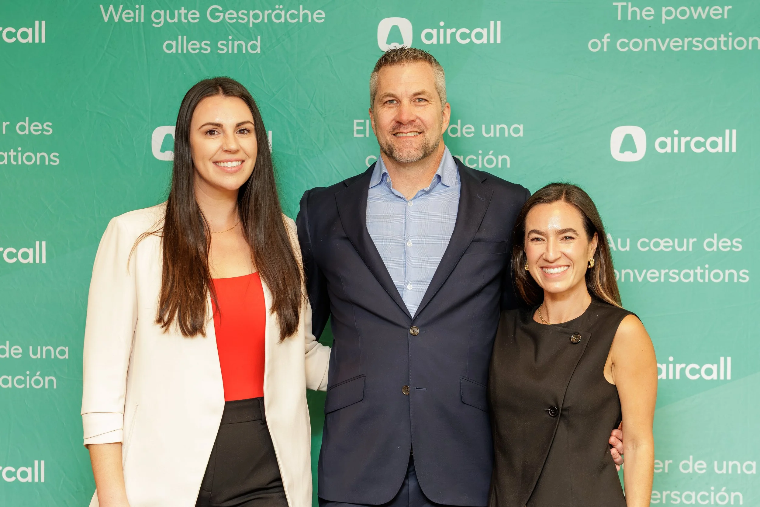 Three professionally dressed individuals standing together and smiling in front of a green backdrop with white text and logos, including 'aircall' and text in multiple languages.