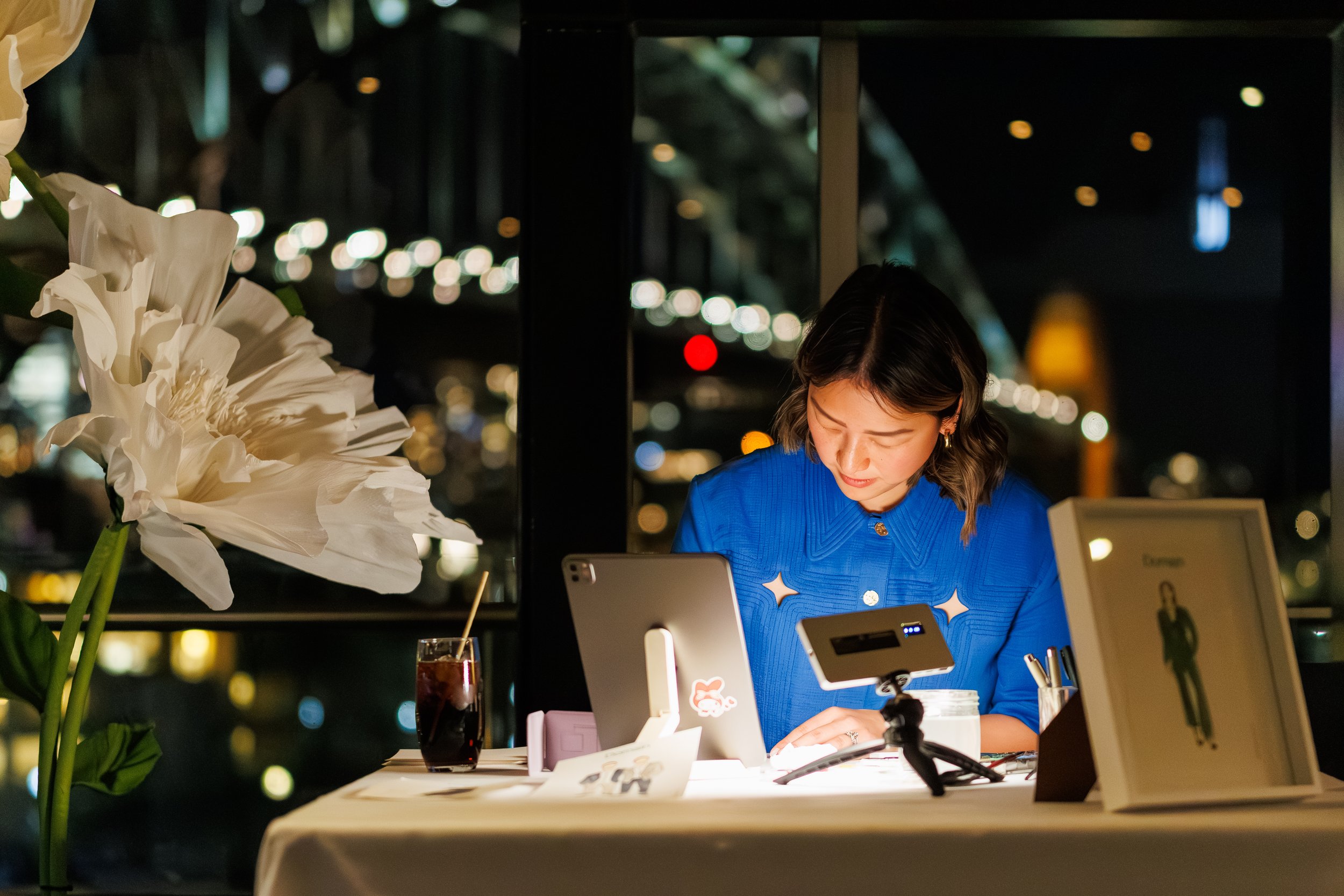 A woman wearing a blue top is working at a counter with a tablet device, a glass of dark soda with ice and a straw in front of her. There are bright city lights outside the window behind her, creating a bokeh effect, and a large white flower in the f