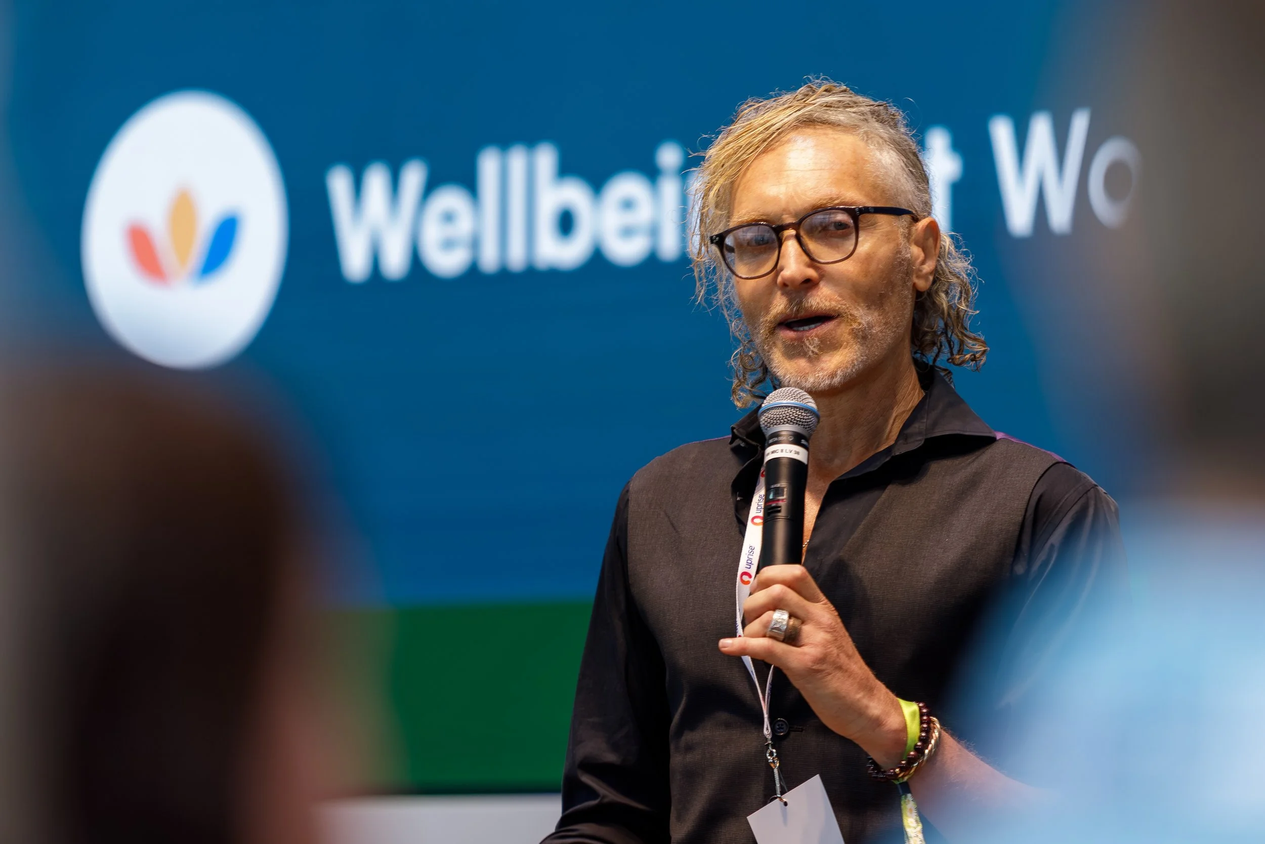 A man with glasses and long curly blonde hair speaking into a microphone at a conference, with a blurred blue background featuring the Welbex logo and text.