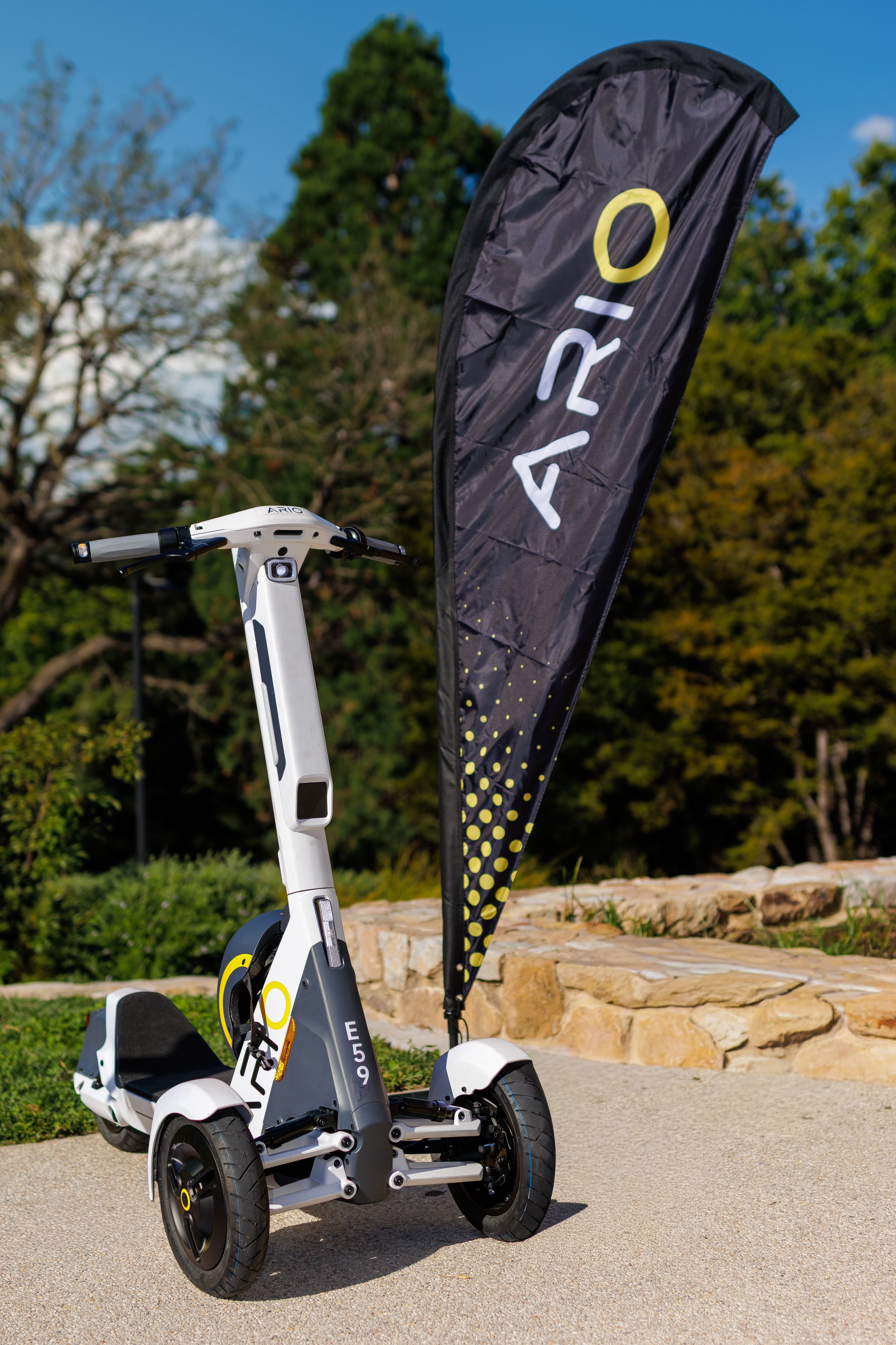 An electric scooter with a black and yellow design, positioned outdoors on a paved surface next to a stone border and green bushes. A black flag with yellow and white lettering is next to it, and trees with autumn foliage are in the background under 