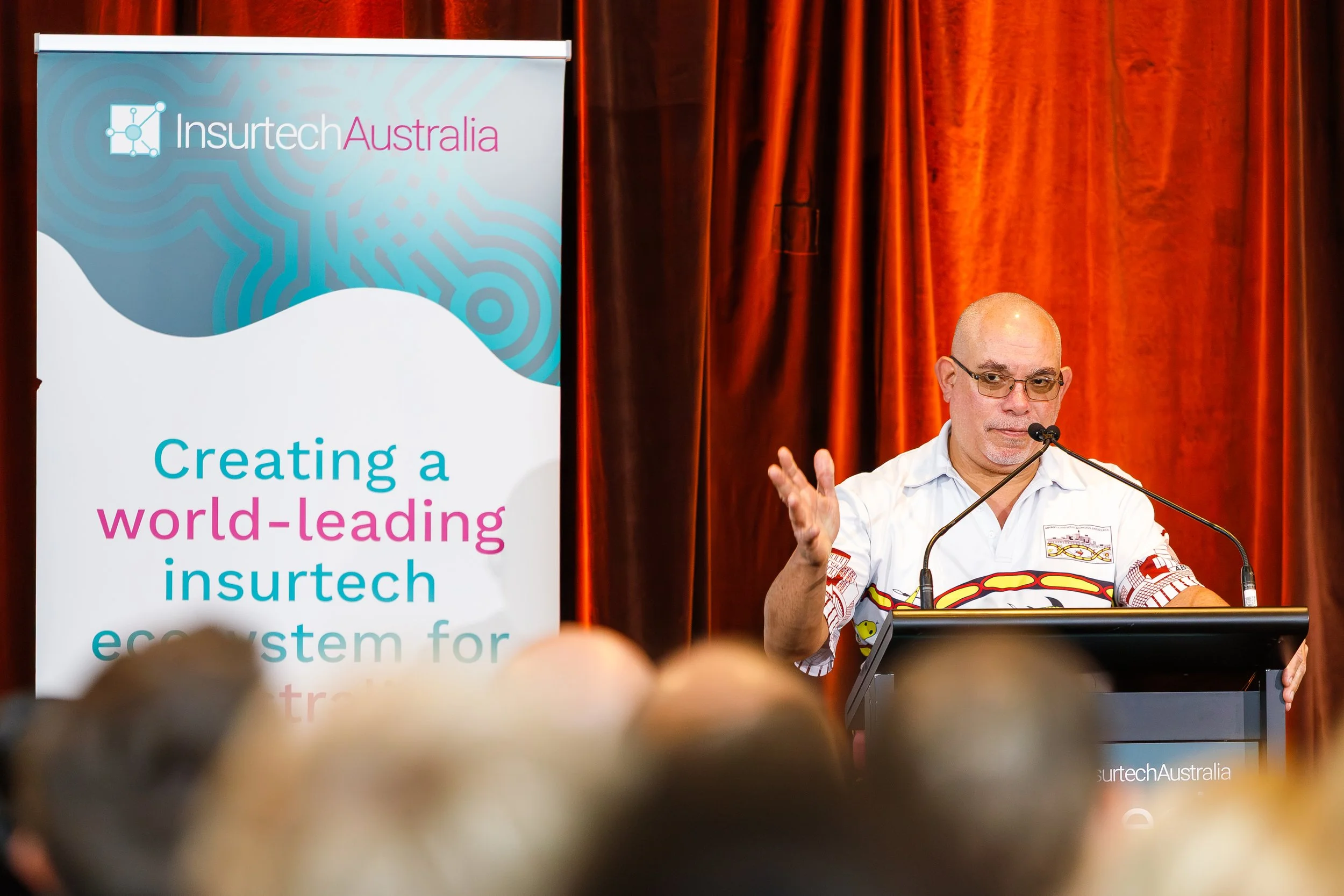 A man with glasses and a bald head giving a presentation at a conference, standing behind a lectern, with an orange curtain in the background. To his left, there's a banner for Insurtech Australia that reads "Creating a world-leading insurtech ecosys