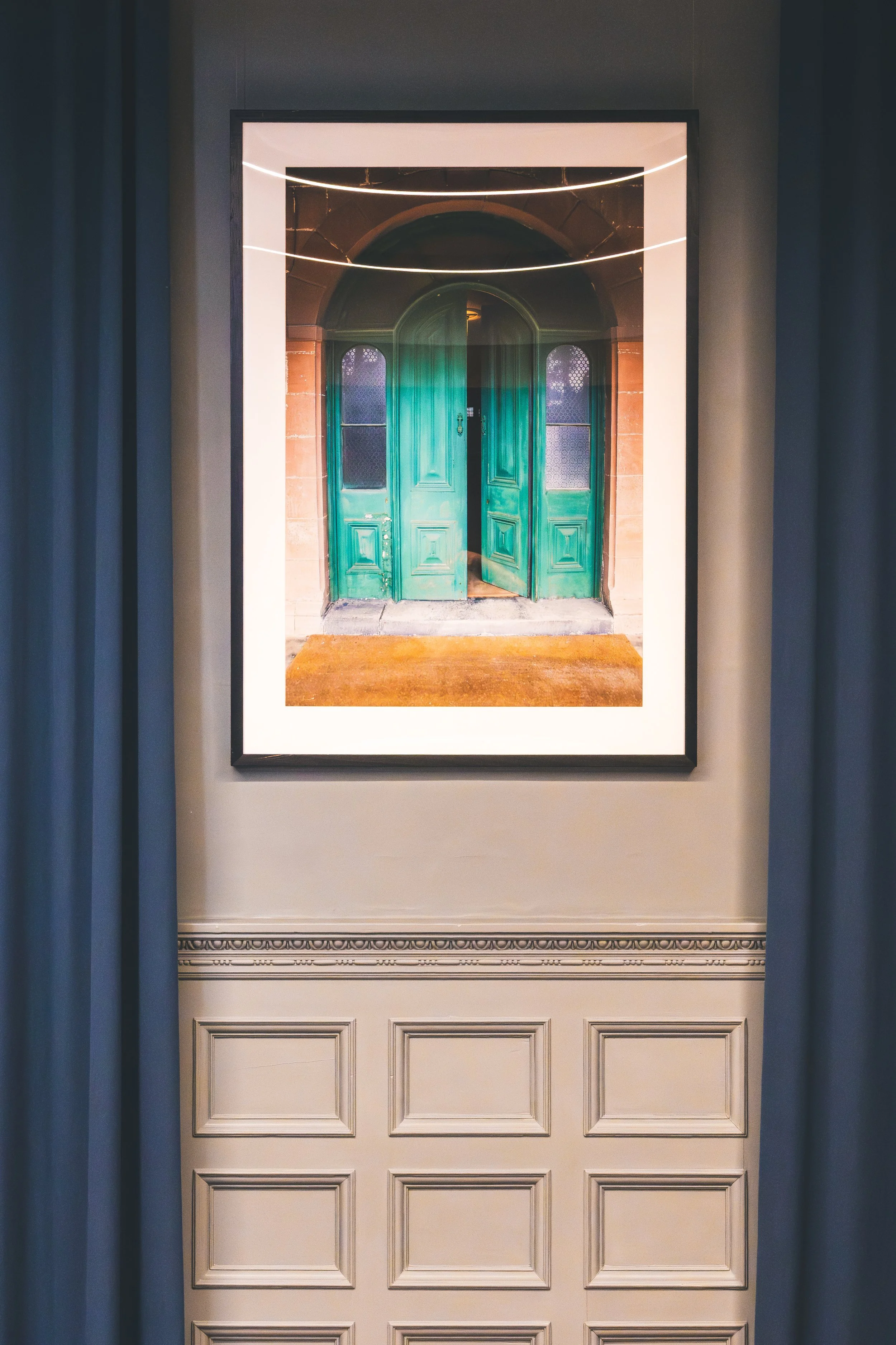 Framed photograph of a green arched double door with glass panels, mounted on a wall with curtains on either side.
