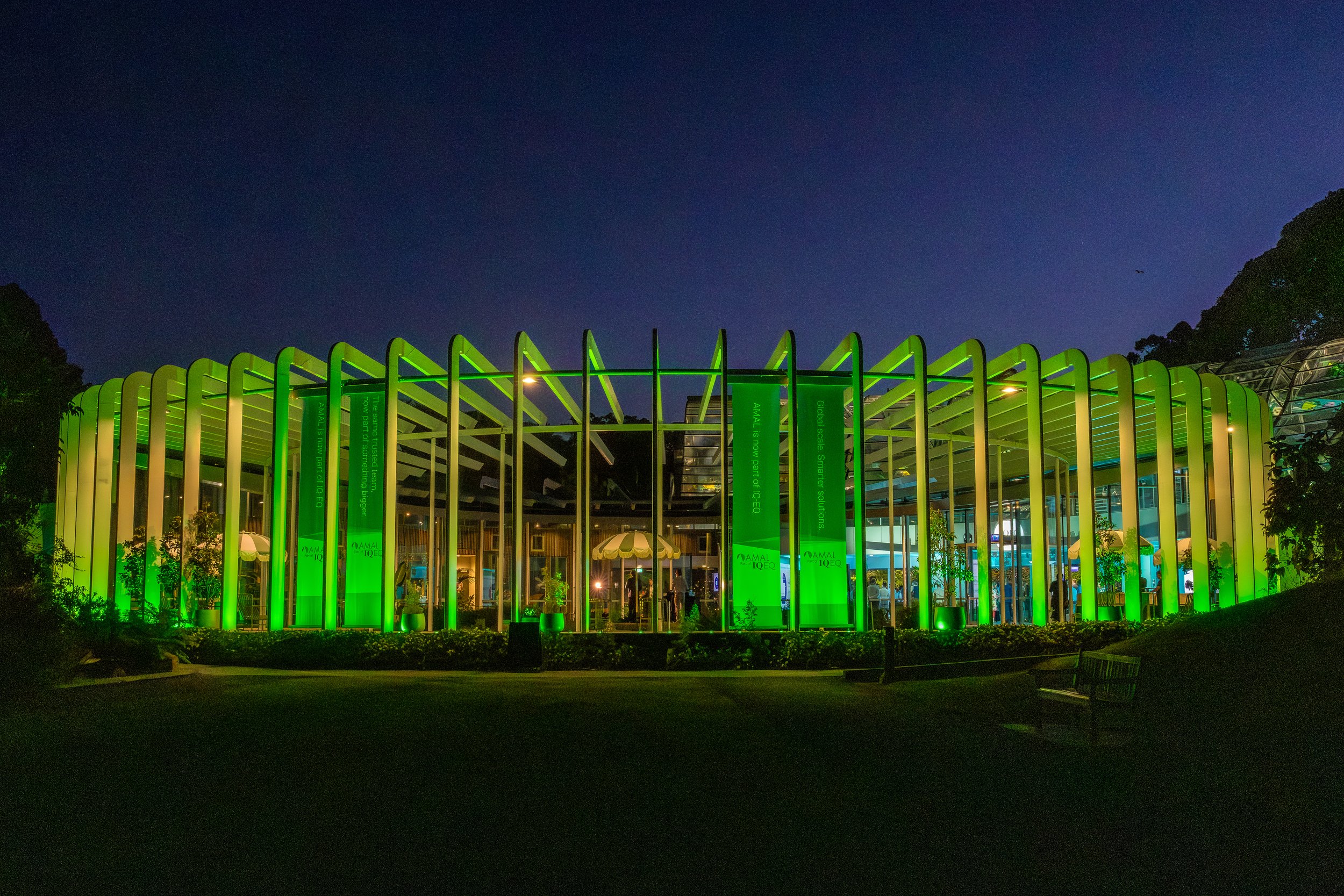 A modern building with a green illuminated exterior at night, featuring vertical arches and glass windows, with trees and a dark sky in the background.