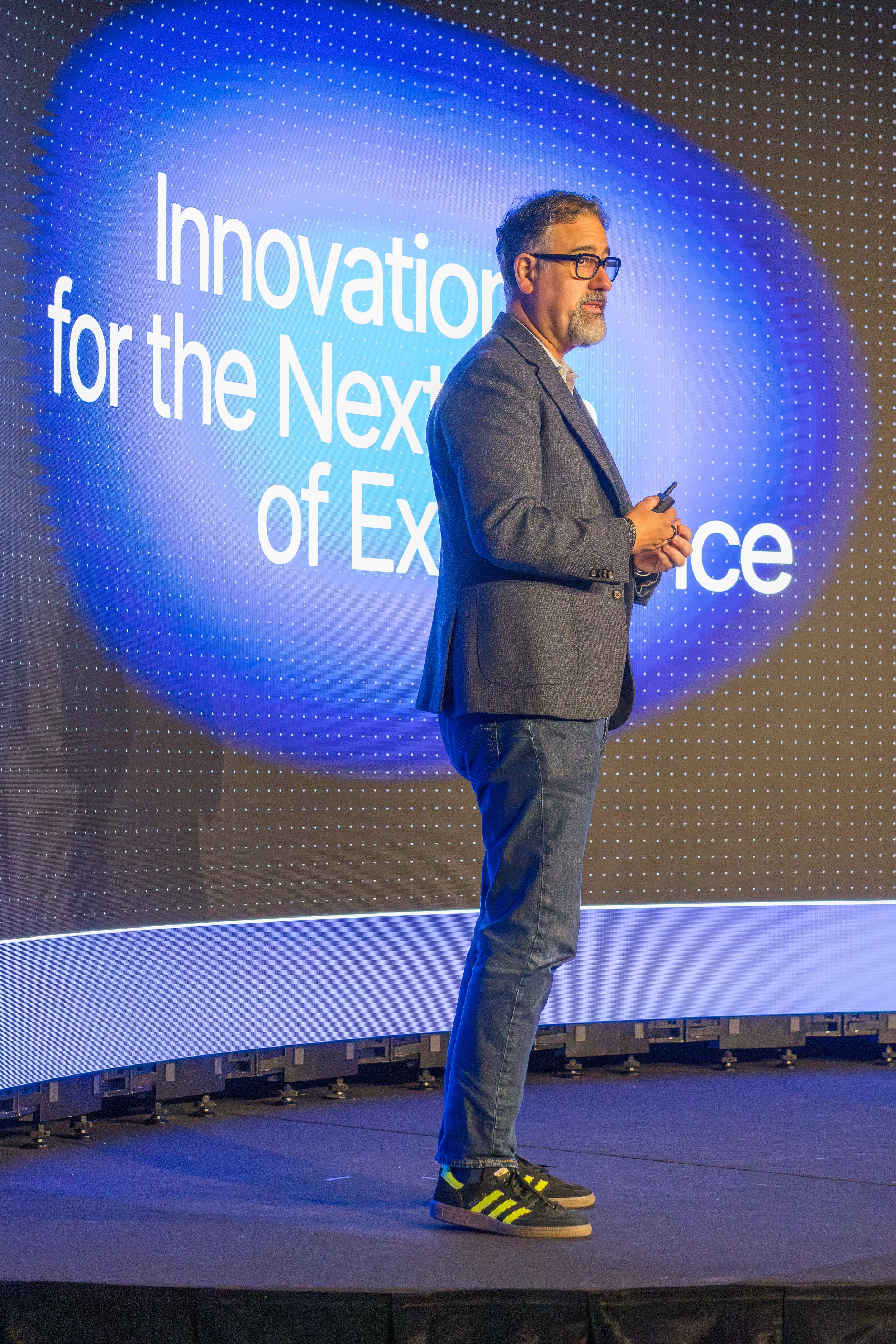 A man with glasses, a beard, and short hair stands on a stage with a dark floor. He is wearing a gray blazer, jeans, and black sneakers with yellow stripes. Behind him, there is a large digital screen displaying the words "Innovation for the Next of 