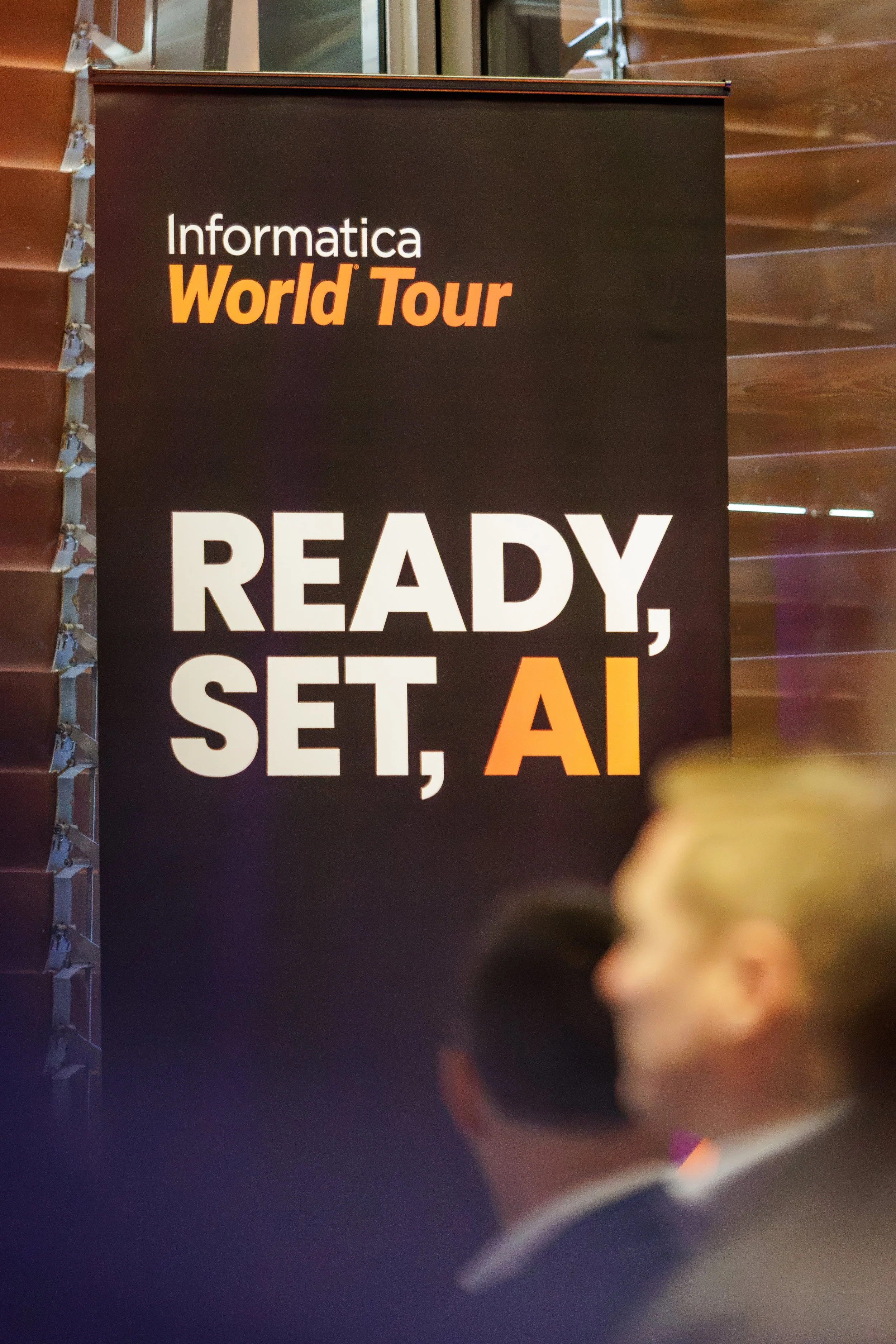 A vertical banner for a technology event titled 'Informatica World Tour,' with the text 'READY, SET, AI' partially visible at the bottom. The banner is black with white and orange text, placed against a background of wooden paneling and glass windows