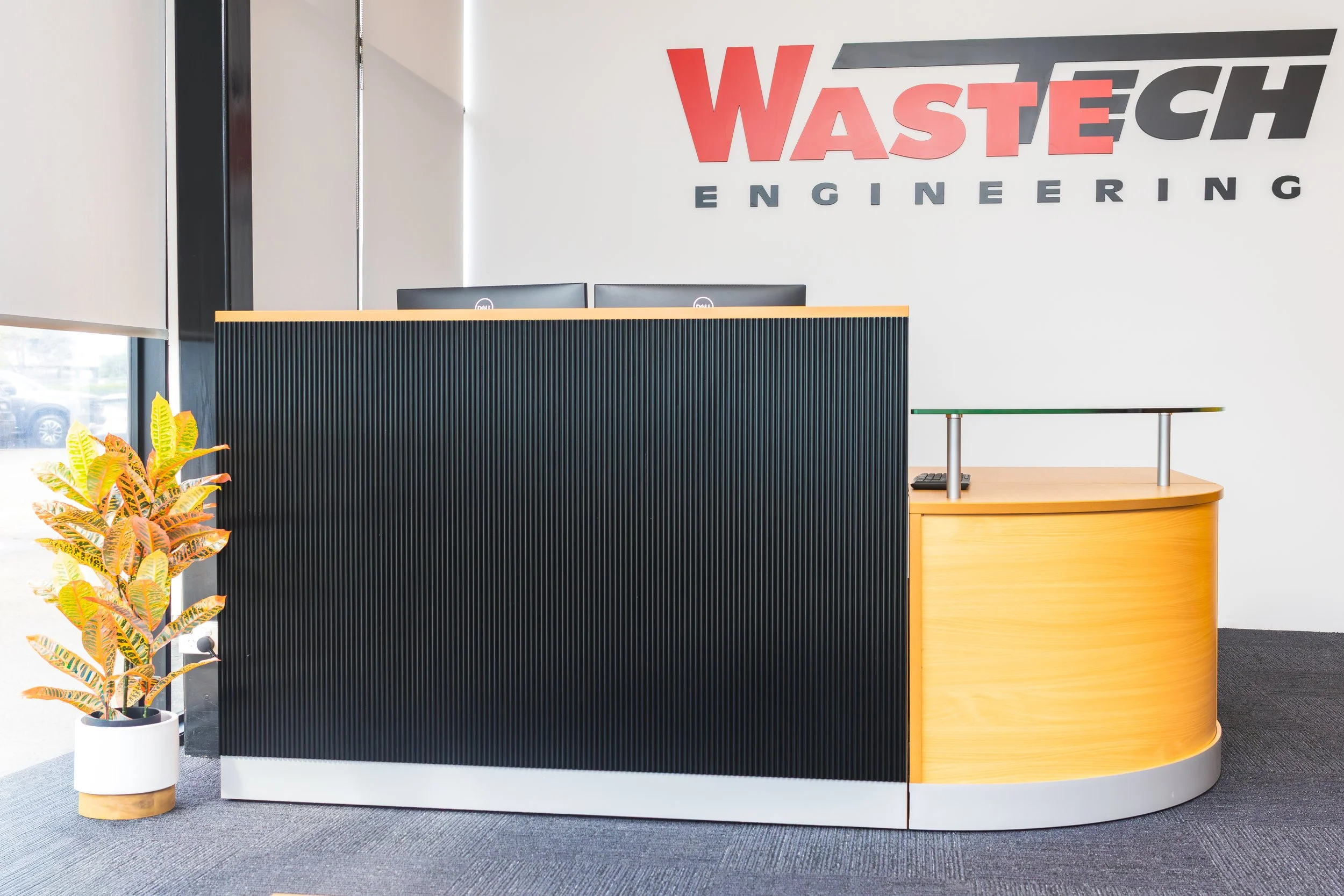 Office reception area with a black and wood curved desk, a potted colorful plant, and a wall sign reading "WASTECH ENGINEERING" in red and black letters.