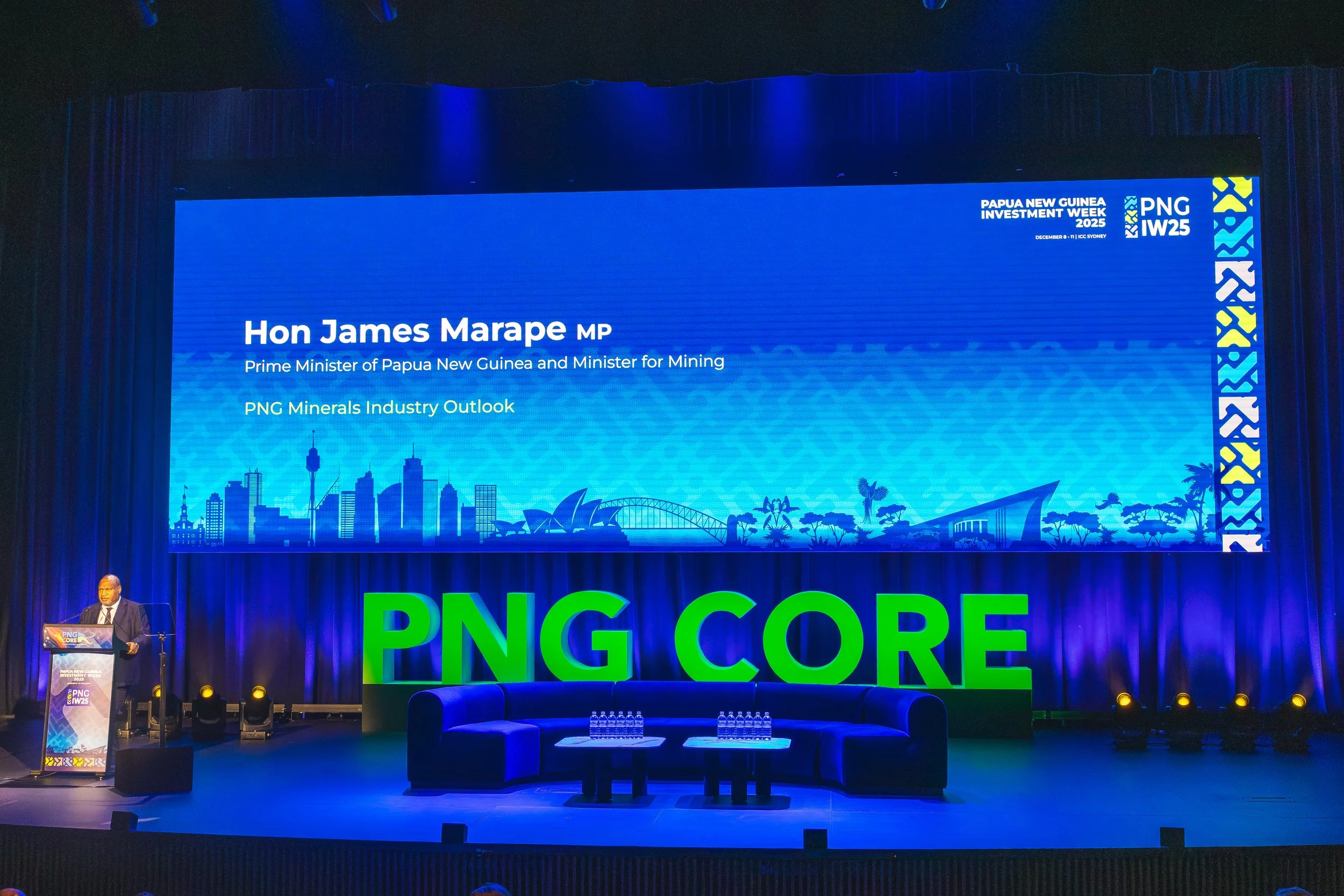 A man at a podium speaking at the Papua New Guinea Investment Week 2025 conference. The large screen behind him displays the event details with the title, date, and location, along with a skyline illustration of Papua New Guinea. The stage is decorat