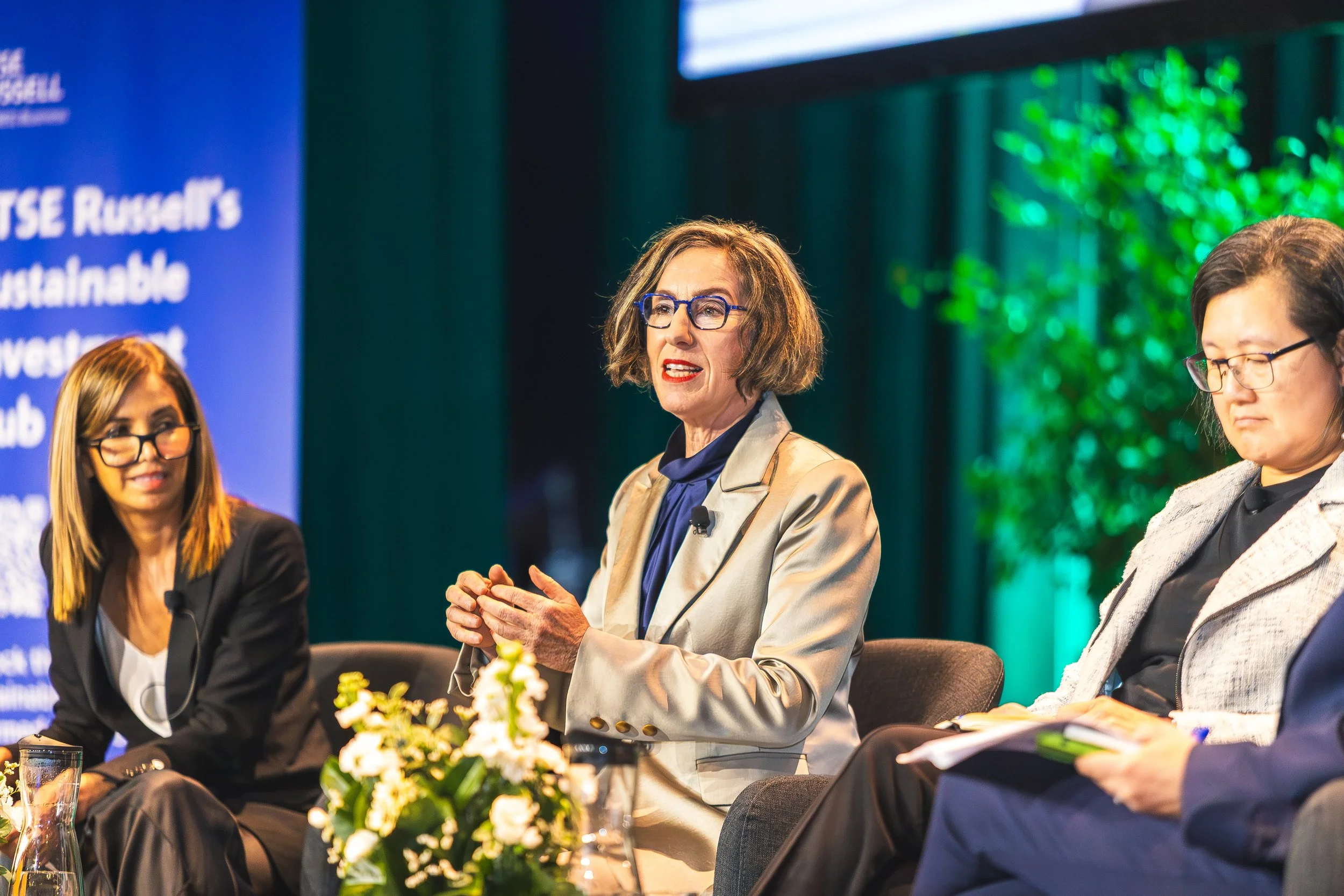 A woman with short brown hair and glasses speaking at a panel discussion, sitting among three other women with conference microphones in front of a blue background with text, and a green decorative tree on stage.