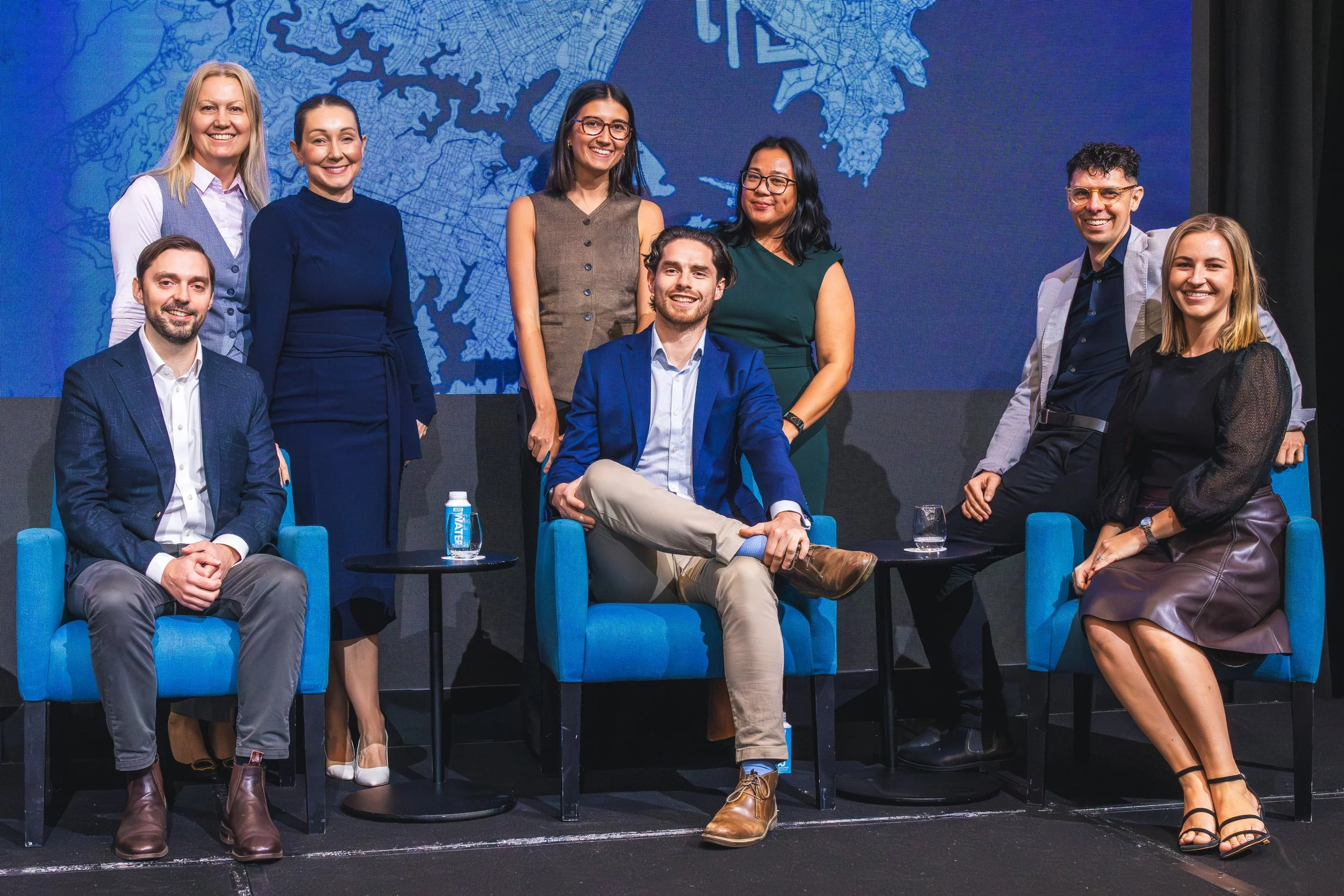 Group of nine diverse individuals at a panel discussion, some seated and some standing in front of a blue background with a city map.