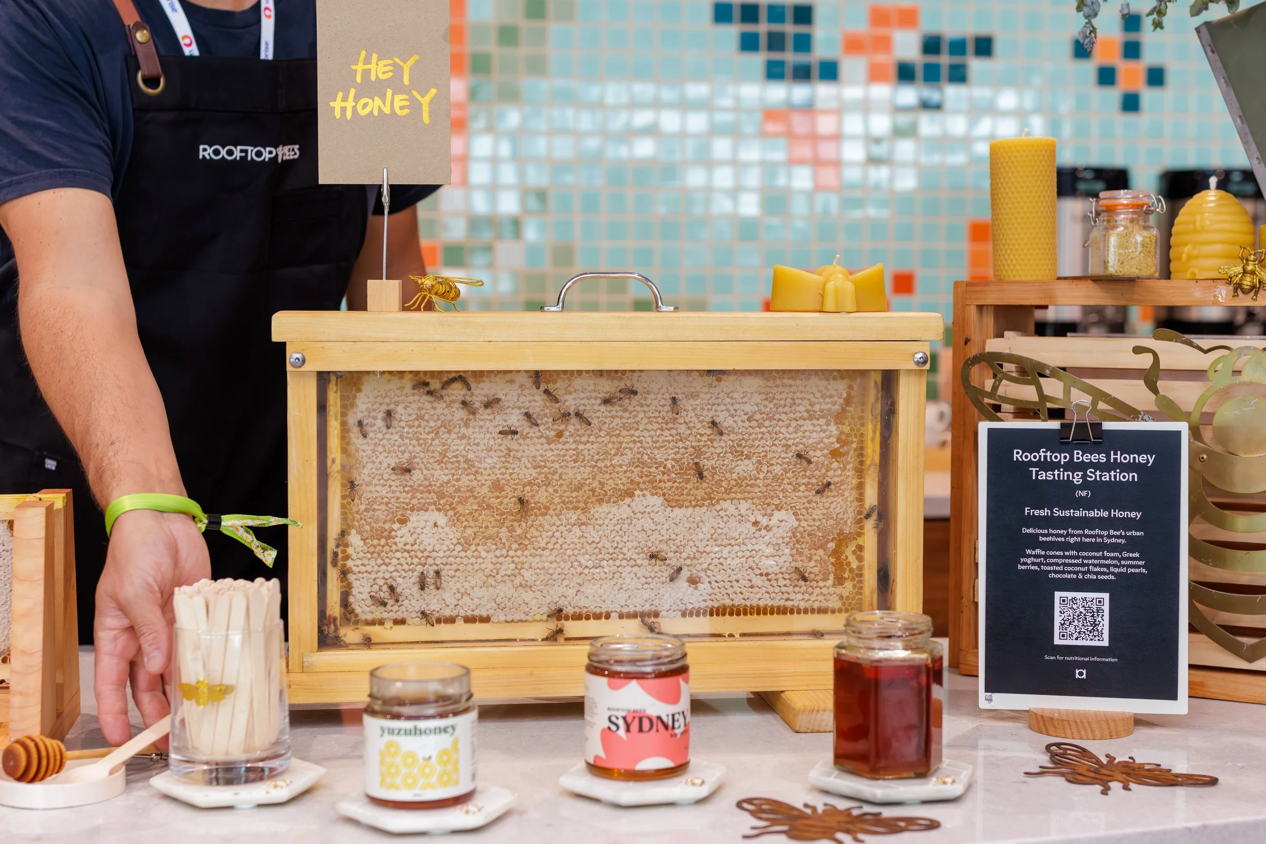 A bee display station with honey jars, honey sticks, and honey-themed products. A person wearing a black apron labeled 'ROOFTOP' stands behind the table. A sign reads 'Hey Honey,' and a larger sign promotes rooftop bees honey tasting, featuring honey
