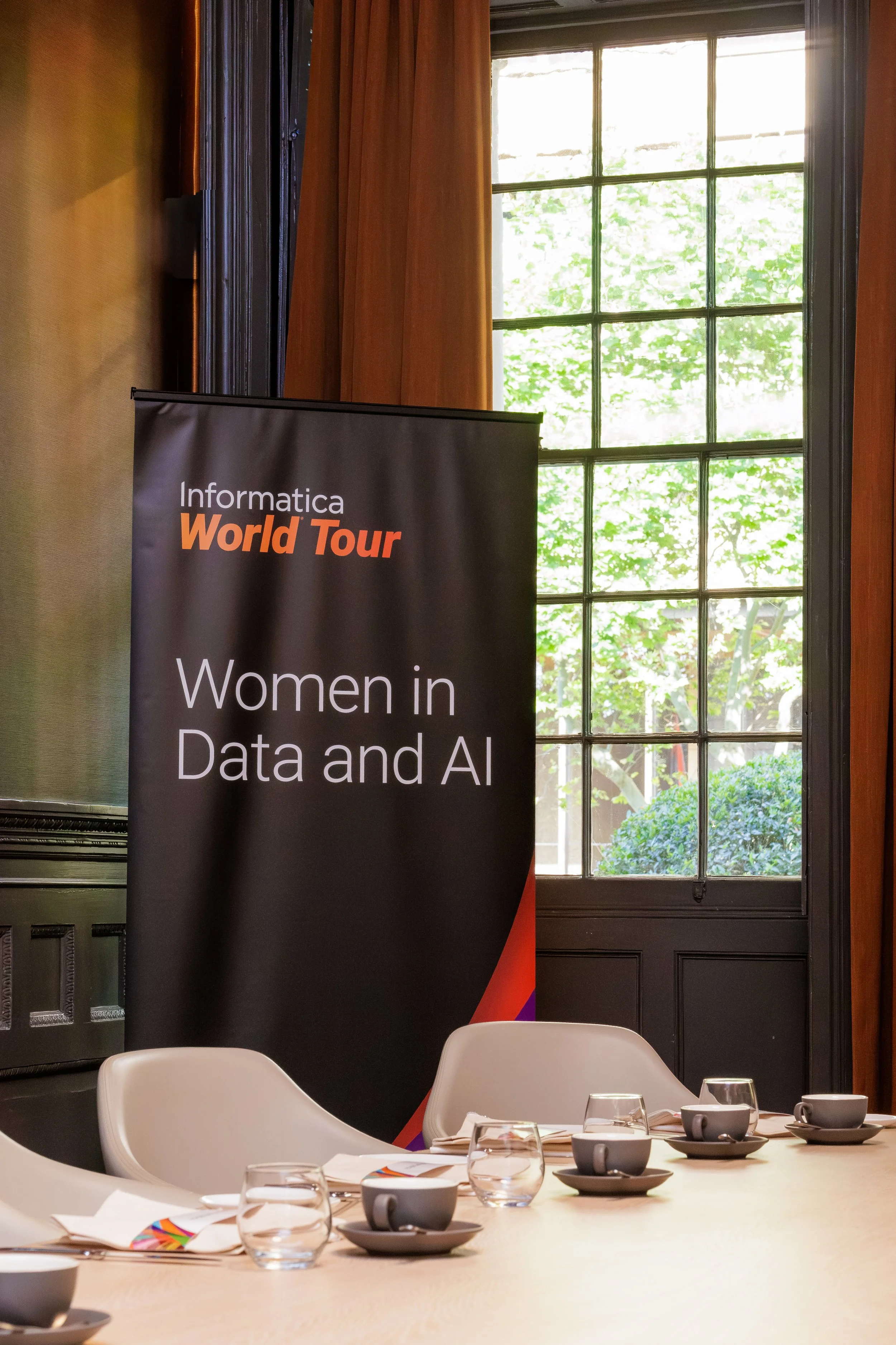 Conference room setup with white chairs, glasses, and cups on a large table. A banner on the side reads 'Informatica World Tour' and 'Women in Data and AI.' Large window behind the banner shows green trees outside.