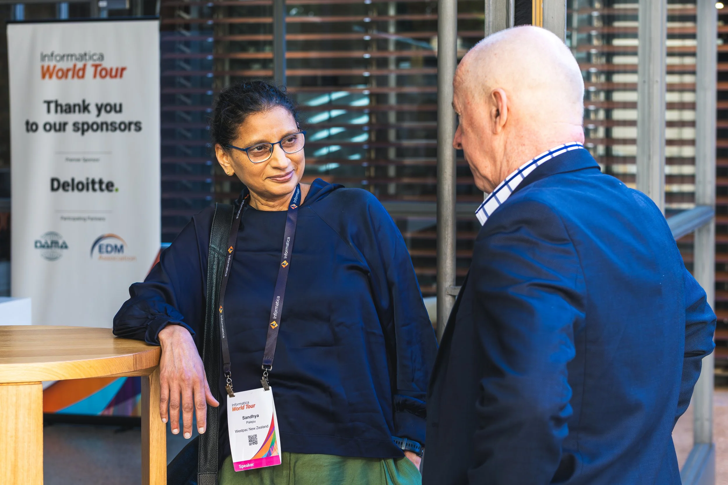 Two people engaged in conversation at a professional event, with a sign in the background that reads "Informatica World Tour." One person is a woman wearing glasses, a navy blue top, and a conference badge, and the other is a man with a bald head, we