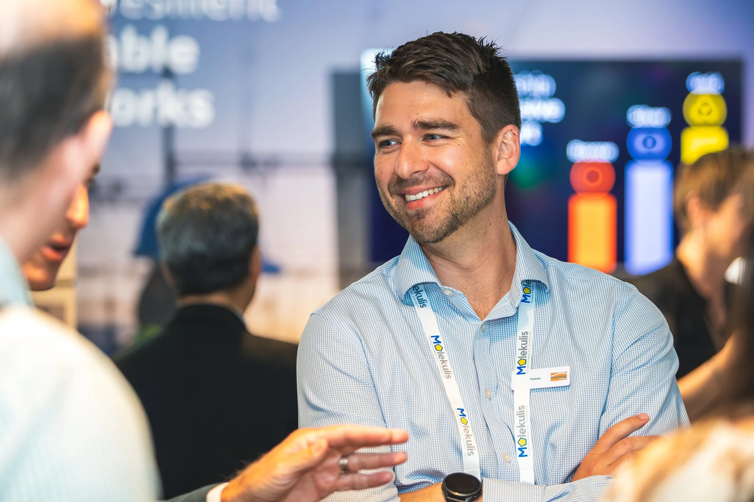 A man with a beard smiling and wearing a blue checkered shirt and a lanyard with the logo 'ekukulis' engaging in conversation at what appears to be a professional event or conference, with screens and other people in the background.